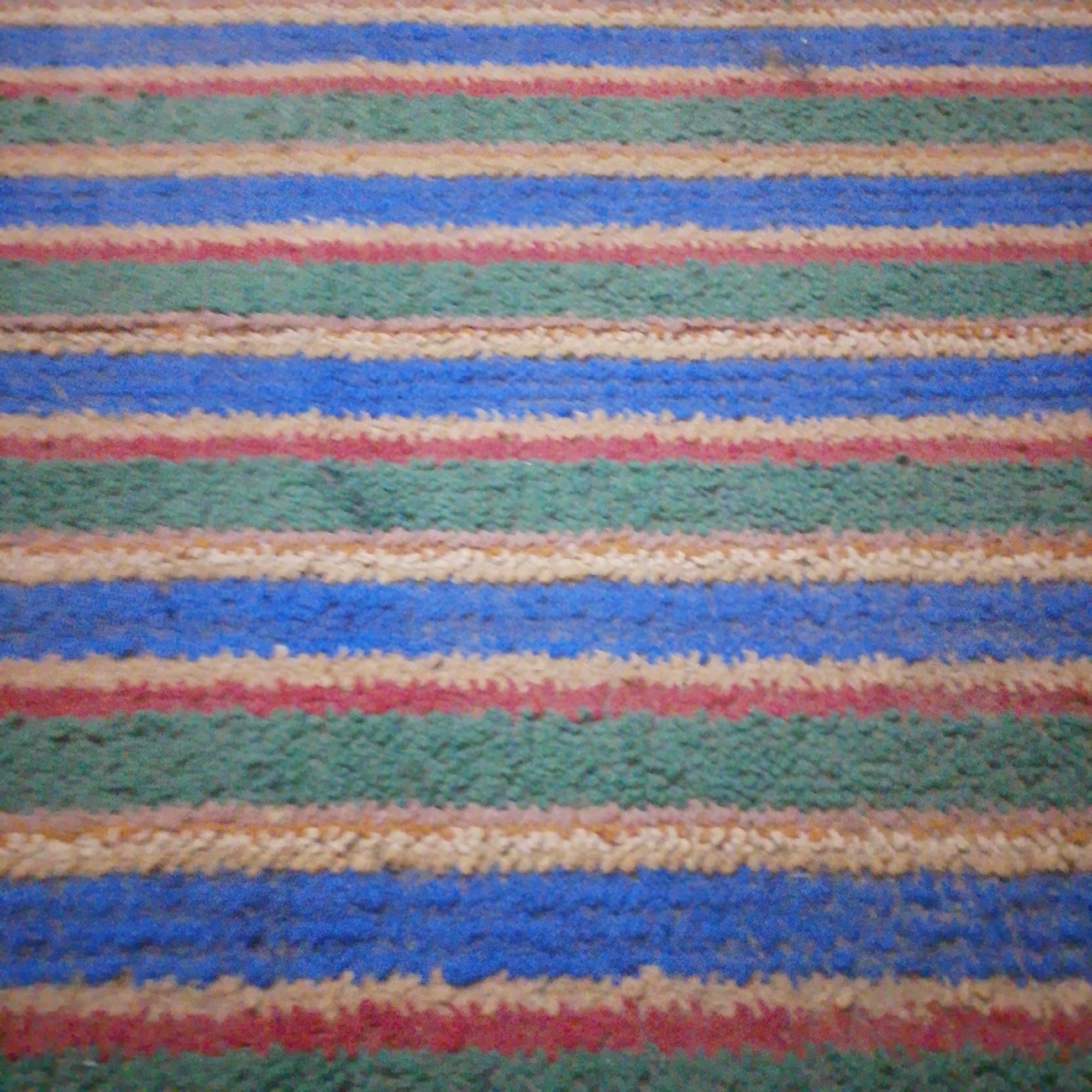 The image shows a close-up, horizontally-oriented view of a carpet with a striped pattern. The stripes are of varying widths and colors, including blue, green, red, and cream. The texture of the carpet appears to be looped or cut pile. There are no people, objects, or discernible actions visible. The scene is a close-up of a carpet and provides no information about its location in Maiburiji, Nigeria, or any other city.