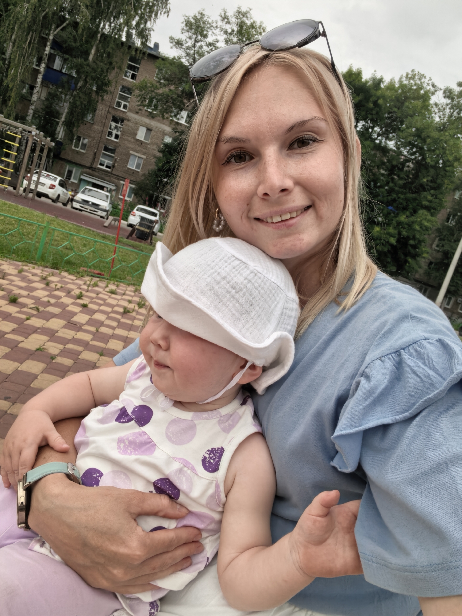 In Sterlitamak, Russia, a blonde woman wearing sunglasses atop her head and a blue shirt smiles at the camera while holding a baby. The baby, wearing a white hat and a white dress with purple polka dots, looks to the side. They are sitting on a patterned ground, possibly a park or playground area, with a residential building and trees in the background. There are cars parked on a street behind a green fence.