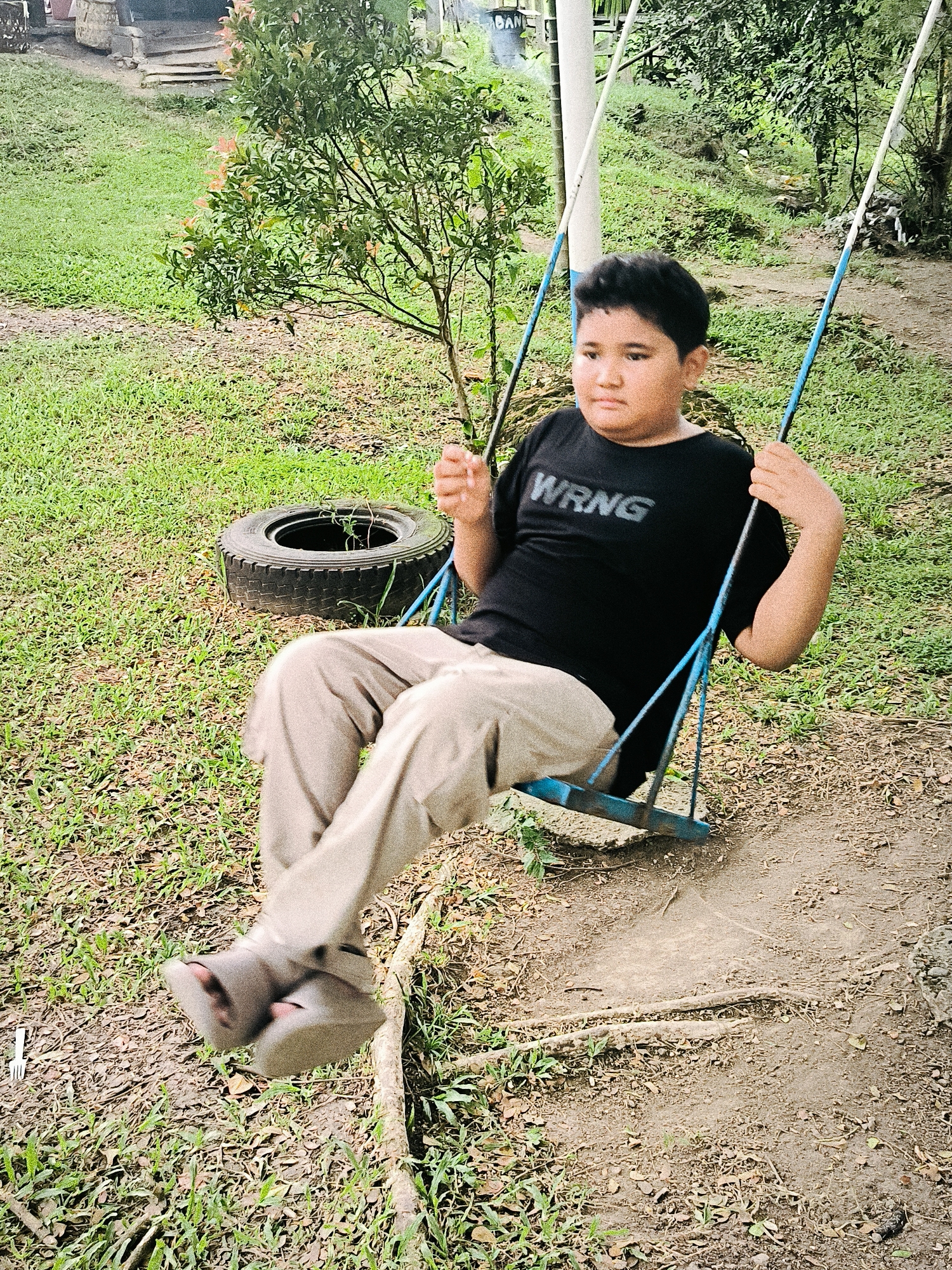 A young boy is captured in motion on a swing in an outdoor, green setting in Lambarotunong, Indonesia. The boy, who appears to be of elementary school age with dark hair, is positioned in the center-right of the frame, mid-swing. He wears a black short-sleeved t-shirt with the white text "WRNG" prominently displayed across the chest, and light tan or beige trousers. His feet, clad in sandals, are blurred due to the motion of the swing. His expression is neutral, looking slightly off-camera to the right.
The swing itself is simple, with blue-painted metal bars and white ropes or chains. The ground beneath the swing is a worn dirt path, surrounded by short, green grass interspersed with patches of dry leaves and exposed roots. To the boy's left, an old car tire lies on the grass, seemingly repurposed or discarded, with some small plant growth inside. Behind him and to his right, there are various trees and lush green foliage, indicating a natural, somewhat unkempt environment, possibly a park, backyard, or rural area. The lighting suggests it is daytime, with soft, diffused light, possibly overcast. In the background, hints of structures or buildings can be seen through the trees, further suggesting a semi-developed outdoor space.