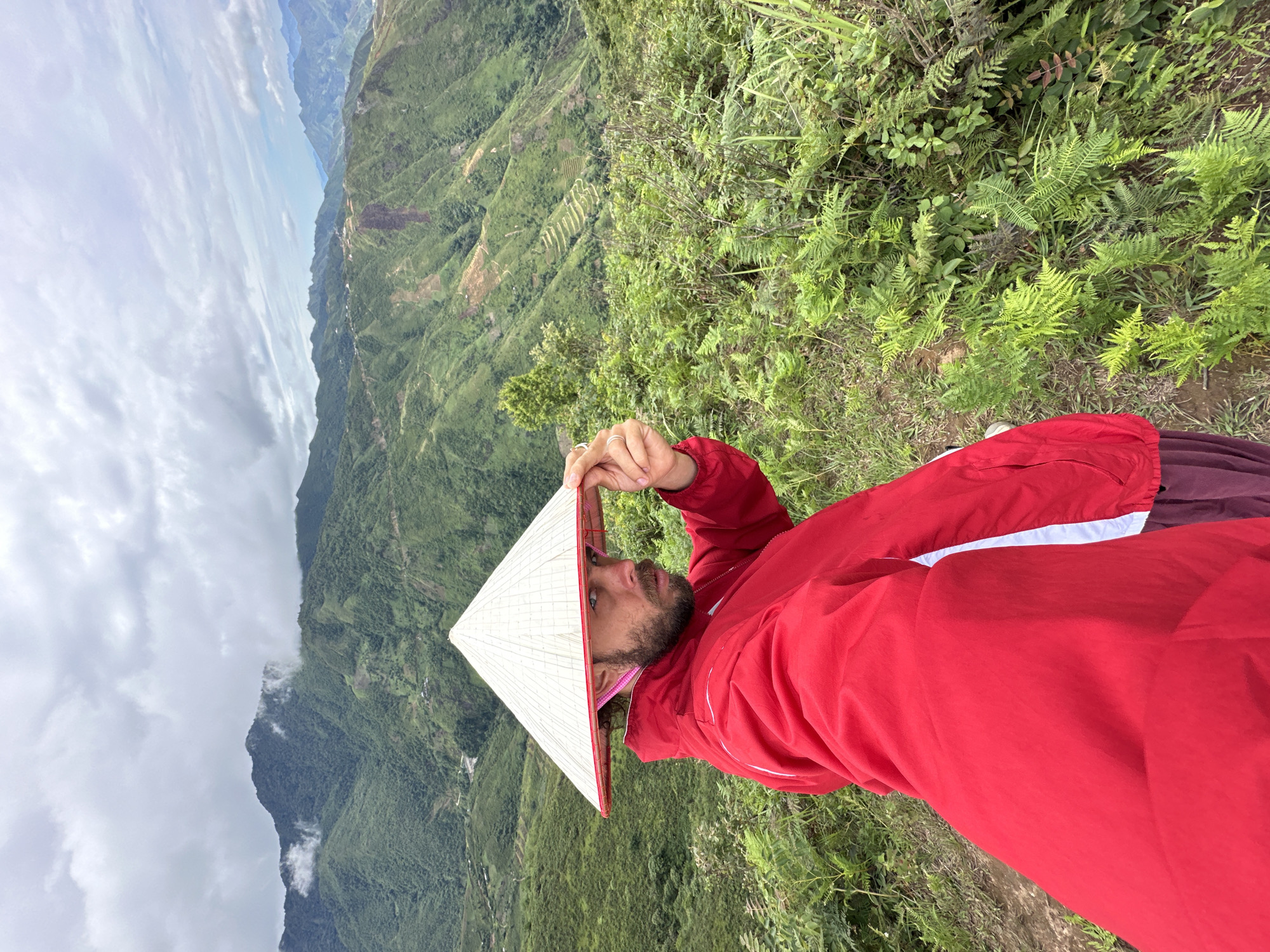 A man wearing a conical hat and a red jacket poses for a selfie on a mountain in Tà Xùa, Vietnam. The man's left hand is holding the brim of the hat. The background shows a vast landscape of lush green mountains with terraced fields and a cloudy sky. The man has a beard and is looking at the camera. He is standing on a grassy slope with ferns and other vegetation.