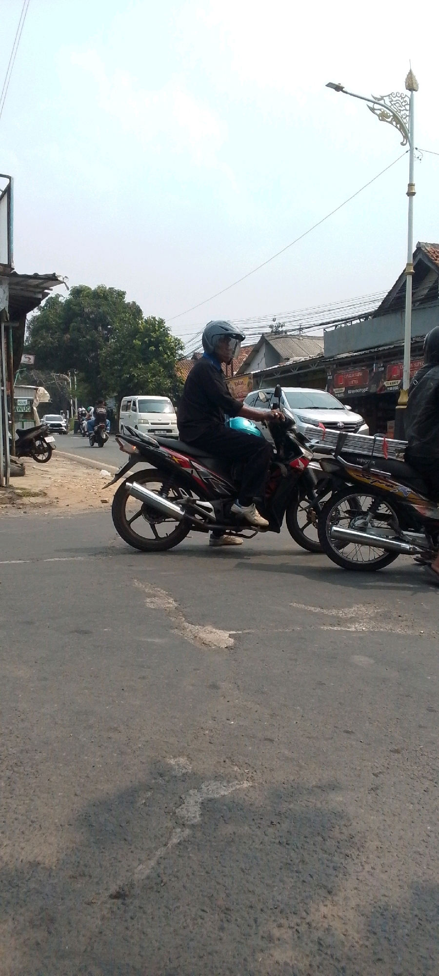 The image captures a street scene in Cilaku, Indonesia, where an elderly man, wearing a blue helmet and a dark long-sleeved shirt, rides a black and red motorcycle. He is seen from the side, looking forward, with a clear visor on his helmet. The motorcycle has a chrome exhaust pipe and visible decals. Behind him, another motorcycle is partially visible, also with a rider. The street is paved with asphalt and shows signs of wear, including a patch of lighter-colored material and some cracks. In the background, there are buildings with corrugated metal roofs, utility poles with numerous wires, and a streetlight with an ornate design. Several other vehicles, including a white van and a silver car, are present on the road, indicating moderate traffic. Trees with dense foliage are visible in the distance, and the sky is overcast with some clouds. The overall impression is a typical day in a moderately busy Indonesian town.