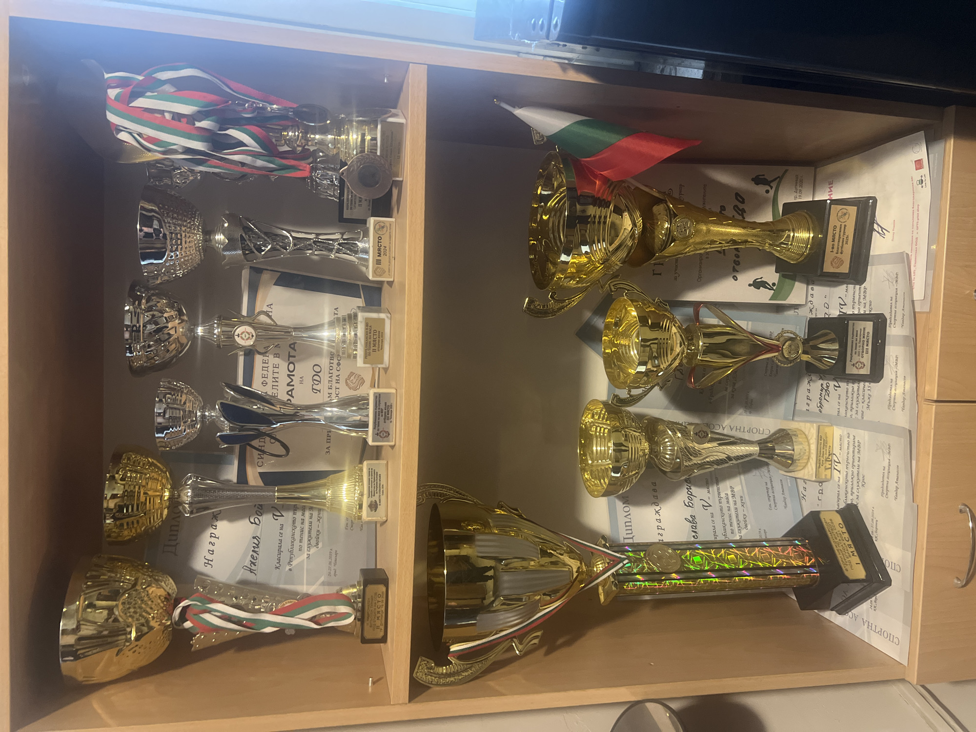 The image displays a collection of sports trophies and awards neatly arranged on wooden shelves within an indoor setting, likely in Sofia, Bulgaria. The trophies are predominantly gold-colored, with some silver accents, and vary in size and design. Many are decorated with ribbons featuring the national colors of Bulgaria (white, green, and red). A small Bulgarian flag on a stick is also prominently placed among the awards on the right shelf.
Behind and beneath the trophies, several certificates and diplomas are visible, confirming the nature of the achievements. The awards signify success in various sports, indicated by phrases such as "I МЯСТО" (1st Place), "II МЯСТО" (2nd Place), and "III МЯСТО" (3rd Place) written on the plaques and bases. Visible Bulgarian text mentions specific competitions including "по тенис на маса" (table tennis) for "служители на МВР" (employees of the Ministry of Interior) in "двойки - жени" (women's doubles), with one event dated "24-27.06.2019 г. град Чепеларе." Other referenced events include a "Благотворителен турнир" (Charity Tournament) for "футбол" (football) from "19.09.2020г.", "Републиканско първенство" (Republican Championship) in "приложно ориентиране" (applied orienteering) and "Крос" (cross-country running) for "Мъже 35+" (Men 35+), and "В ОТБОРНО ЖЕНИ" (In Teams, Women). Names like "Анелия Бой..." and "Чавдар Атанасов," noted as "Председател на Спортна асоциация" (Chairman of a Sports Association), are also discernible on some documents. The display reflects significant sporting accomplishments.