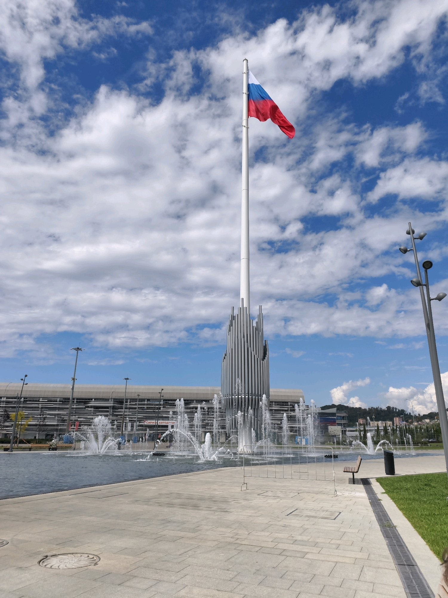 The image depicts a wide-angle view of a plaza with a large fountain in the foreground and a prominent flagpole with the Russian flag flying against a bright blue sky with scattered white clouds. The flagpole is situated behind the fountain, and to its right, a tall, modern building with a ribbed facade is visible. In the background, a long, linear building with a dark, horizontally slatted exterior is prominent, likely a part of the Sirius federal territory's infrastructure.
The plaza area is paved with light-colored rectangular stones, and a section of it features a circular manhole cover. The fountain consists of multiple jets of water spraying upwards and outwards into a large, dark-colored basin. To the right of the fountain, a wooden bench sits on the paved area, and a black trash receptacle is nearby. Beyond the plaza, a well-maintained strip of green grass is visible.
The scene is set in the city of Sirius, Russia, a federal territory known for its scientific, educational, and sporting facilities. The architectural style suggests a contemporary urban design, consistent with the development in Sirius. The lighting suggests a sunny day, likely mid-morning or afternoon. No individuals are clearly discernible within the frame, though faint figures might be present in the distant background. The overall atmosphere is one of public space, cleanliness, and civic pride, underscored by the presence of the national flag.