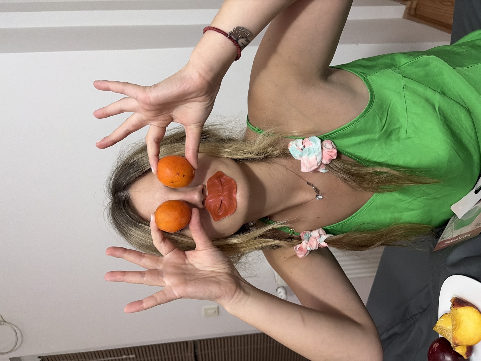 In Budva, Montenegro, a young woman with blonde hair in pigtails, adorned with colorful scrunchies, is captured in a close-up shot. She is wearing a vibrant green tank top and has a playful expression, holding two small, round, orange fruits up to her eyes, resembling a face. A lip-shaped patch is affixed to her mouth. Her right arm displays a beaded bracelet with a heart-shaped pendant. A plate with sliced peaches and plums sits to her left, and a white tissue packet is visible on the surface in front of her. The background is a plain white wall.