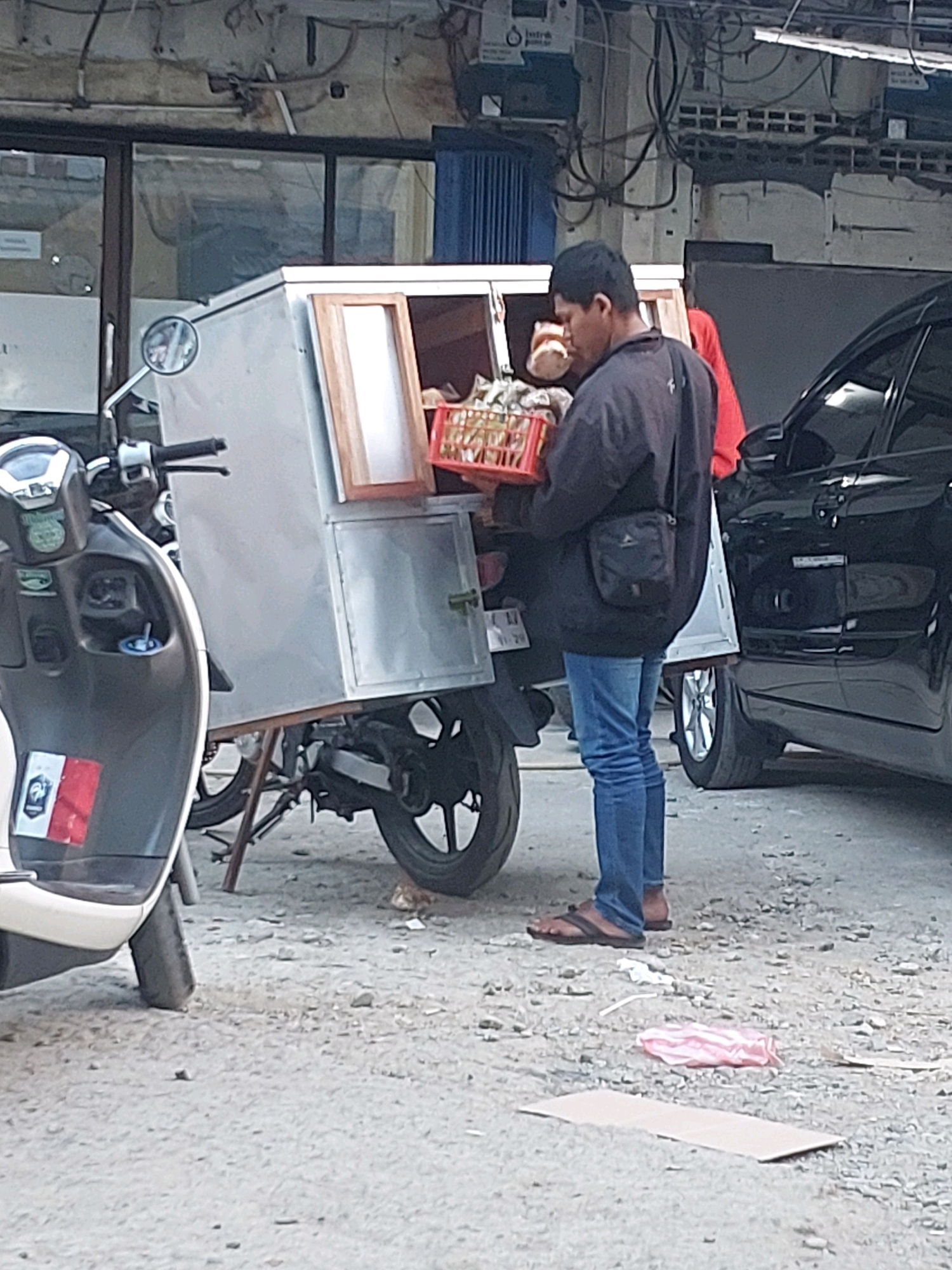 The image depicts a street scene in Ambon, Indonesia, where a man is standing next to a food cart mounted on a motorcycle. The man, wearing a dark jacket, blue jeans, and flip-flops, is holding a red basket filled with food items. The food cart is silver and has a wooden framed window. A black car is parked behind the food cart. In the foreground, a white and grey scooter is partially visible. The ground is unpaved and covered with gravel. The background shows a building with an open front and various wires and pipes on its exterior.