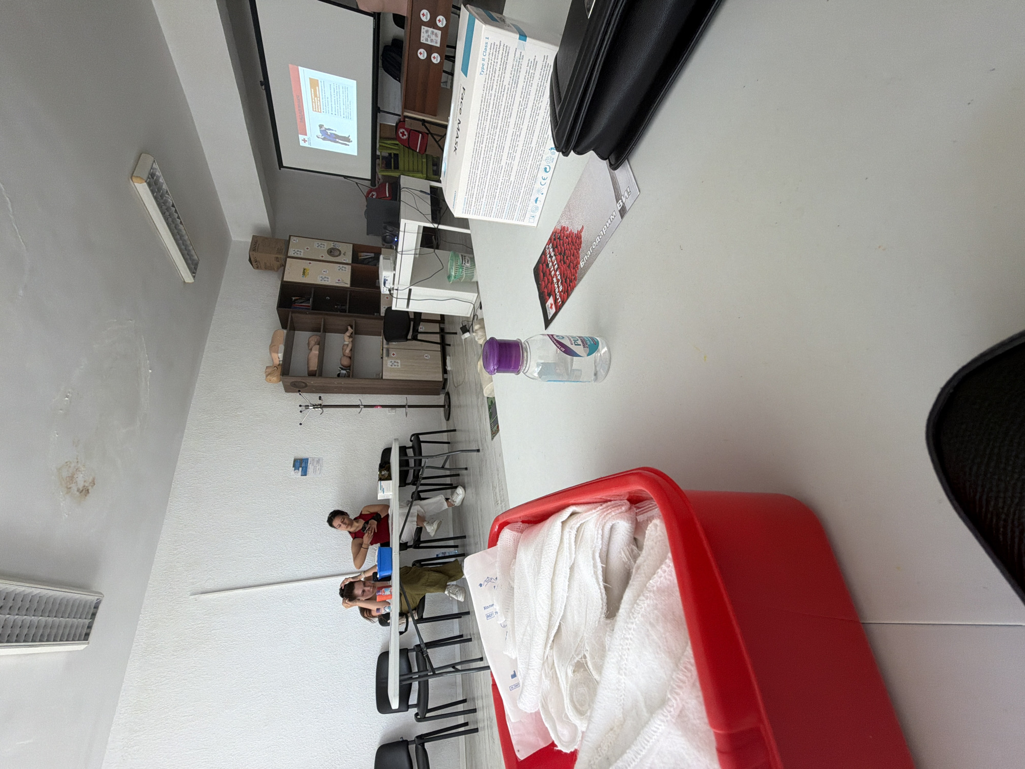 The image depicts a classroom or training room setting. In the foreground, a bright red plastic container holds white folded cloths, possibly bandages or medical supplies. To the right of the container, a small bottle of hand sanitizer with a purple cap sits on a white table. Further to the right, a box labeled "Face Mask" and a black clutch bag are also on the table. In the background, a projector screen displays an informational slide with images and text, suggesting a presentation or lesson. Two women are seated at a table in the mid-ground, facing away from the camera. One woman, wearing a red top, appears to be looking at her phone or an object in her hand. The other woman, wearing a green top, has her hands on her head and is looking down. The room has white walls and a tiled ceiling with fluorescent lighting fixtures. Shelving units are visible on the left side of the room, containing what appear to be medical training dummies. The overall scene suggests a medical training session or a first-aid course is in progress or has recently concluded. The location is identified as Burgas, Bulgaria, based on the metadata provided.