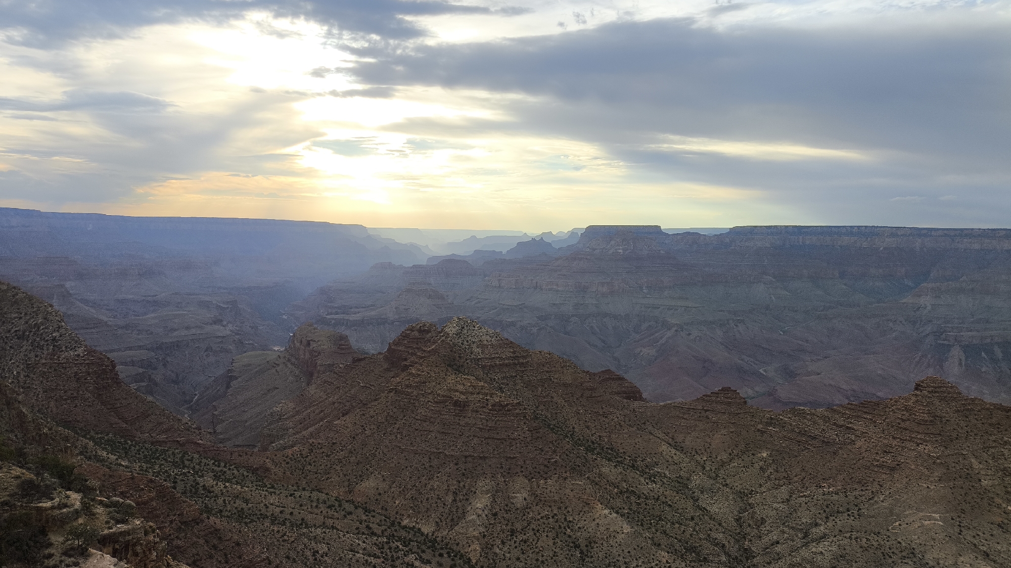 This is a panoramic landscape photograph of the Grand Canyon in Grand Canyon Village, Arizona, United States. The image captures a vast expanse of the canyon, characterized by its layered rock formations, steep cliffs, and deep ravines. The sun is setting or rising, casting a warm, golden light across the sky and illuminating the canyon walls with dramatic shadows and highlights. The sky is filled with a mix of clouds, with some areas of bright sunlight breaking through. The foreground features rugged, rocky terrain with sparse vegetation, leading down into the immense depth of the canyon. In the distance, multiple layers of the canyon are visible, receding into a hazy, atmospheric perspective. The overall impression is one of immense scale, natural beauty, and geological grandeur.