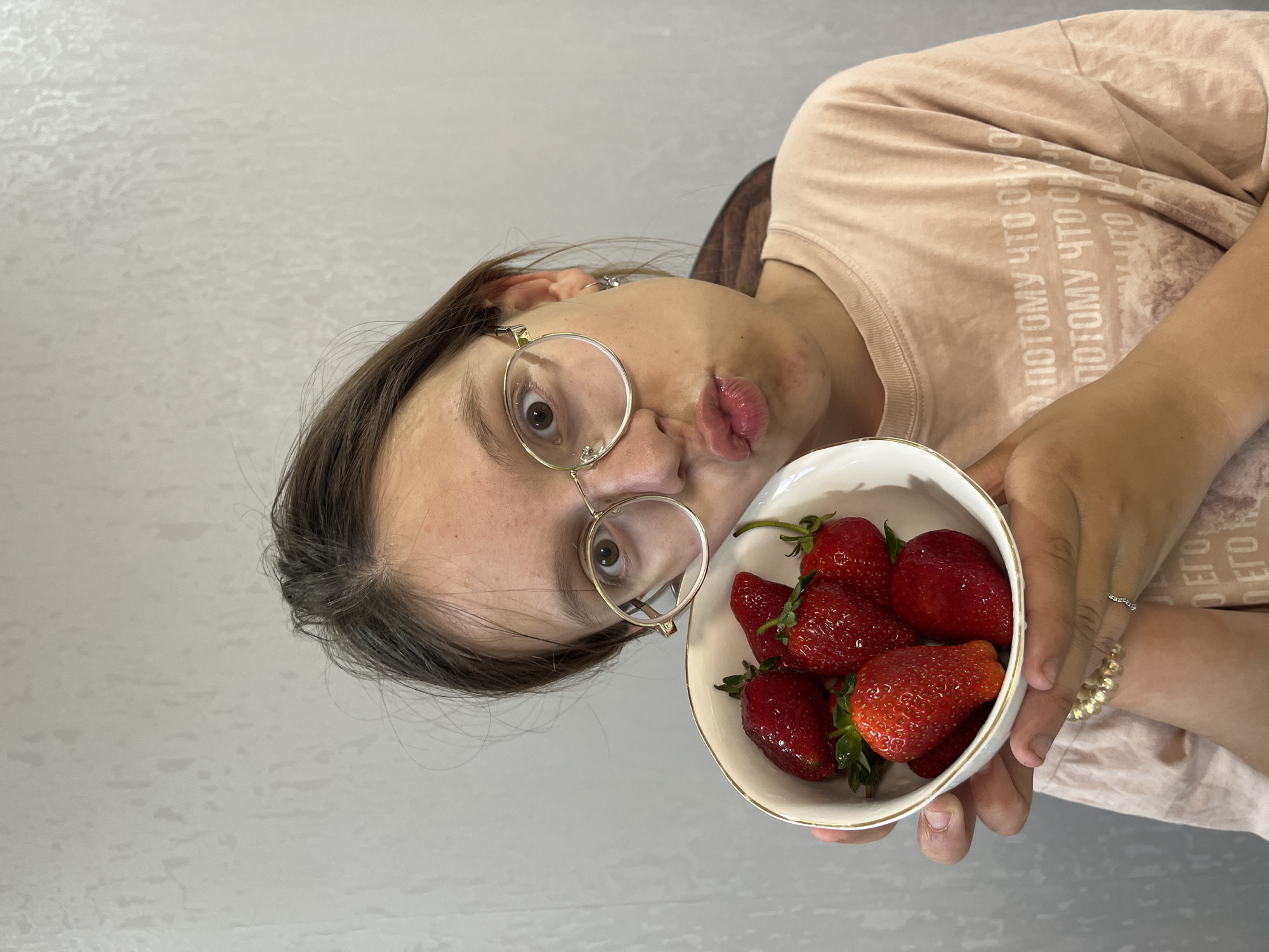 An indoor, eye-level shot captures a young person, possibly female, holding a bowl of strawberries. The individual, with shoulder-length brown hair and wearing round, gold-rimmed glasses, looks directly at the camera with one eye wide open while making a playful "duck face" or puckered lips expression. They are wearing a light peach or pink short-sleeved t-shirt.
In their hands, which are adorned with a simple silver ring on one finger and a gold-beaded bracelet on the wrist, they hold a small, white ceramic bowl with a subtle gold rim. The bowl is filled with several fresh, bright red strawberries, some with their green stems still attached.
The background is a plain, light-colored wall with a subtle textured pattern, suggesting an indoor, possibly domestic, setting. Visible text on the t-shirt in Cyrillic script repeats the phrase "ПОТОМУ ЧТО ОН" and "ПОТОМУ ЧТО", which translates to "BECAUSE HE" or "BECAUSE IT IS." The overall mood conveyed by the expression and fresh fruit is lighthearted and cheerful. The photo was taken in Chaikovsky, Russia.