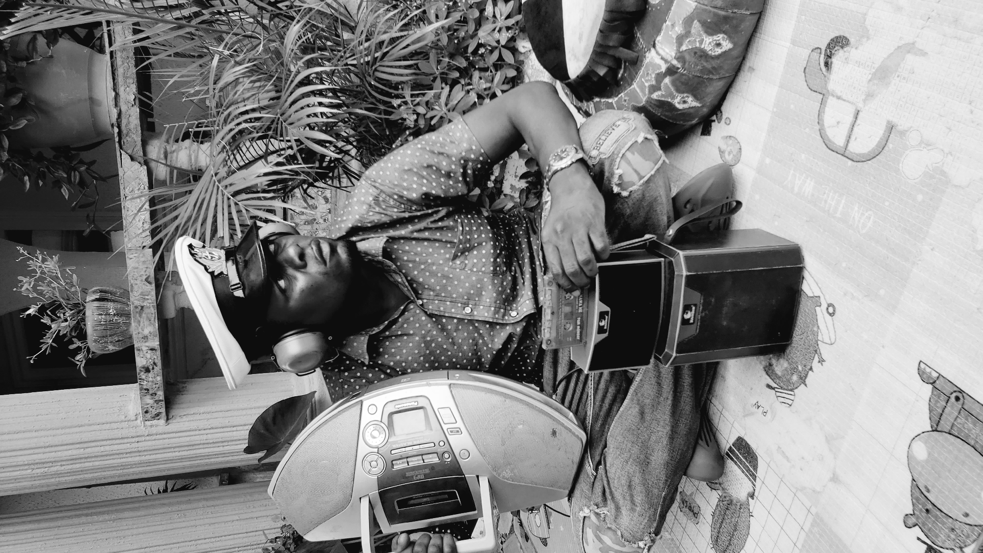 A black and white, medium shot photograph captures a young Black man wearing a dotted short-sleeved shirt, ripped denim jeans, a white captain's hat with a black brim, and headphones. He is seated outdoors on a patterned mat, with lush green foliage and a white pillar in the background, suggesting a residential setting likely in Narayi, Nigeria. The man is holding a portable cassette player and a cassette tape, looking to his right with a thoughtful expression. To his left, a large, oval-shaped boombox sits open, its speaker grill visible. In front of him, a smaller black electronic device, possibly a second cassette player or speaker, is stacked with the cassette tape resting on top. The man's left arm is extended, resting on the boombox, while his right hand holds the cassette tape. His attire, including the ripped jeans and headphones, combined with the electronic devices, suggests a relaxed and perhaps nostalgic mood, possibly related to listening to music. The overall impression is one of a casual outdoor activity.