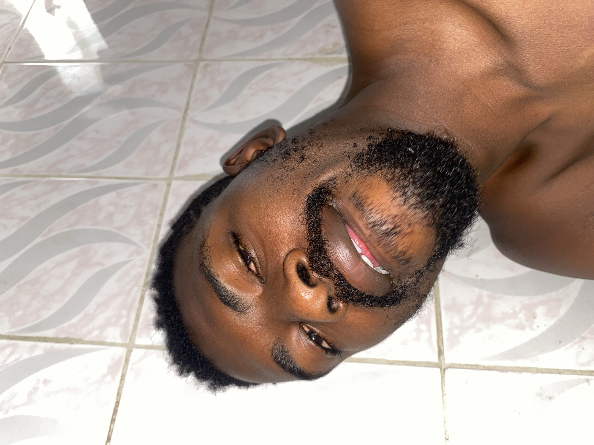 A close-up image shows a young man with dark skin lying on his back on a tiled floor. He has short, dark, curly hair and a well-groomed beard and mustache. His eyes are partially closed, and his mouth is open in a slight smile or grin, revealing his teeth, suggesting a happy or relaxed demeanor. The man's upper body is visible, appearing to be shirtless.
The setting is indoors, featuring light-colored square floor tiles with a subtle, wavy pattern in shades of gray and faint pink, separated by white grout lines. Reflections are visible on the man's skin and the glossy tile surface, indicating an indoor light source. The image is captured from an overhead perspective, looking down at the man. There is no visible text.
