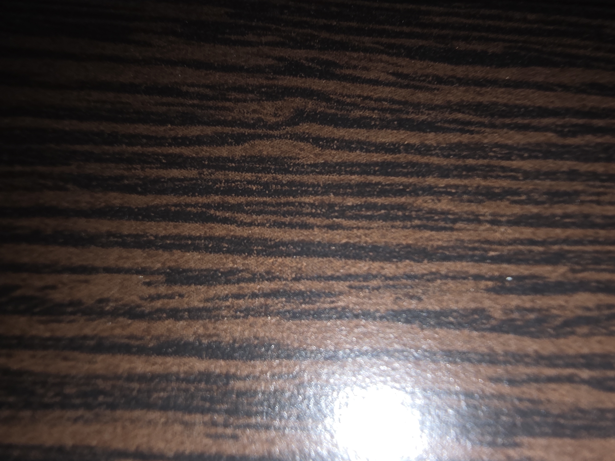 The image shows a close-up, abstract view of a wood-grain pattern. The dominant colors are dark brown and black, with vertical striations that mimic the natural texture of wood. There is a bright, reflective highlight on the left side of the image, suggesting a light source is illuminating the surface. The texture appears somewhat rough and fibrous. The image does not contain any discernible features that would allow for its localization to Ulan-Ude, Russia, or any other specific geographic location within a city. There are no people, identifiable objects, actions, or interactions present.