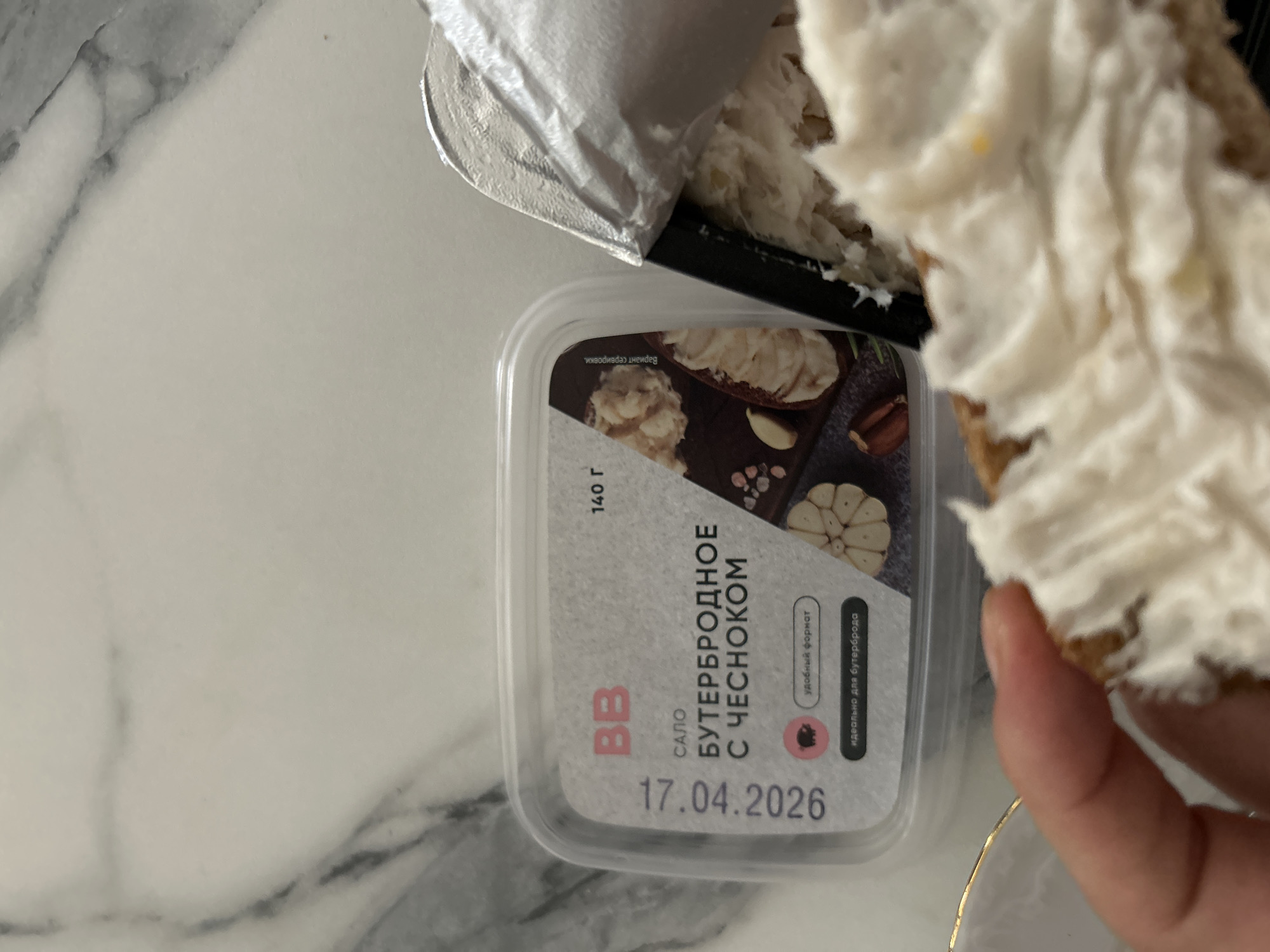 A close-up, slightly overhead shot captures a hand holding a piece of bread generously topped with a white, creamy spread, positioned next to an open plastic container of the same product. The scene unfolds on a white and gray marble-patterned surface, suggesting an indoor setting like a kitchen or dining area in Moscow, Russia.
The clear plastic container features a prominent label in Russian. The main product description reads "САЛО БУТЕРБРОДНОЕ С ЧЕСНОКОМ" (Salo Sandwich Spread with Garlic). Other visible text includes the brand "BB," the weight "140 г" (140 g), and the phrases "удобный формат" (convenient format) and "идеально для бутерброда" (ideal for a sandwich). An expiration date, "17.04.2026," is clearly printed at the bottom of the label. An image on the label illustrates a serving suggestion, showing the spread on bread, accompanied by garlic cloves and spices.
The hand holds a slice of bread covered with the white spread, which has a slightly lumpy texture, consistent with a pork fat-based spread. Silver foil packaging, likely from the container's seal, is partially visible above the container. The activity depicted is the consumption or preparation of a simple meal or snack, highlighting the use of the garlic salo spread on bread. A small portion of a gold-rimmed white dish is visible beneath the hand. The lighting in the image appears to be natural, suggesting it was taken during the daytime.