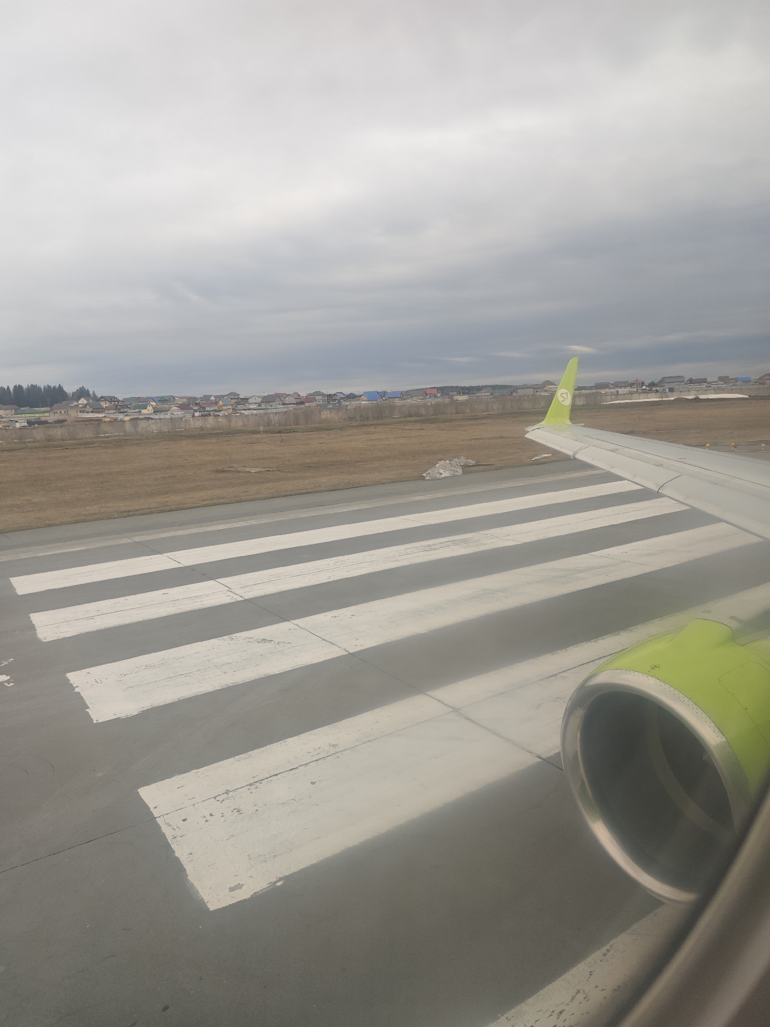 From the perspective of an airplane window, the scene depicts a landing strip in Vaniuki, Russia, marked with parallel white stripes. An S7 Airlines wing and engine are visible in the foreground. Beyond the runway is a flat, grassy field leading to a cluster of residential buildings. The sky is overcast with grey clouds.