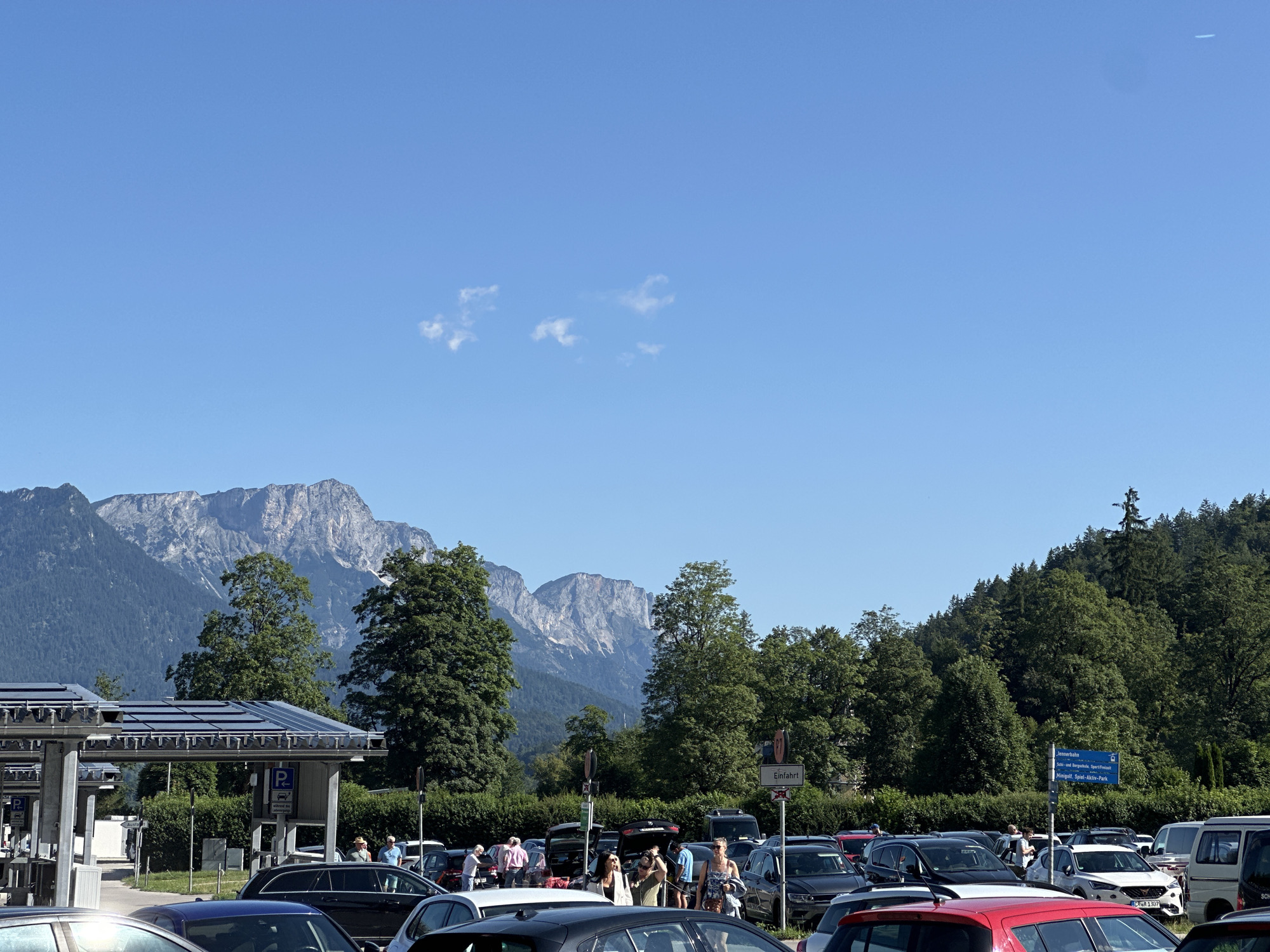 The image depicts a sunny outdoor scene at a parking lot in Schönau am Königssee, Germany. In the foreground, numerous cars are parked, with some people milling about. In the middle ground, there is a structure with solar panels on its roof, likely a covered parking area or charging station. Behind this, a line of lush green trees forms a backdrop, with towering, rugged mountains visible in the distance under a clear blue sky. The overall impression is that of a busy tourist area on a pleasant day.