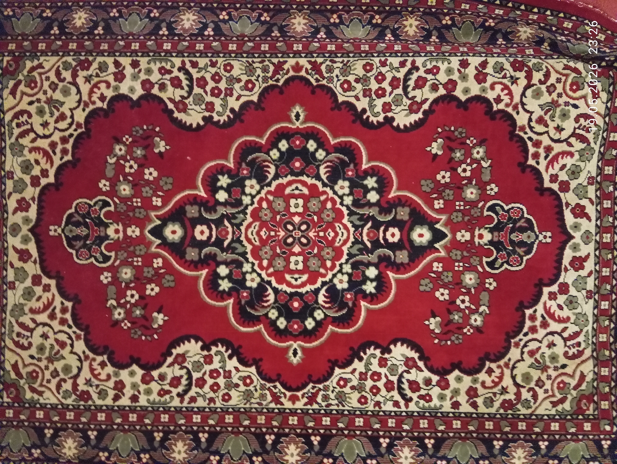 The image shows a red rug with a central, stylized floral medallion design in black, white, and red, surrounded by a red field with scattered floral motifs. The field is bordered by a cream-colored area with smaller floral patterns and an outer border of geometric designs in dark blue, red, and cream. The rug is likely lying flat on a surface. The city and location are unknown.