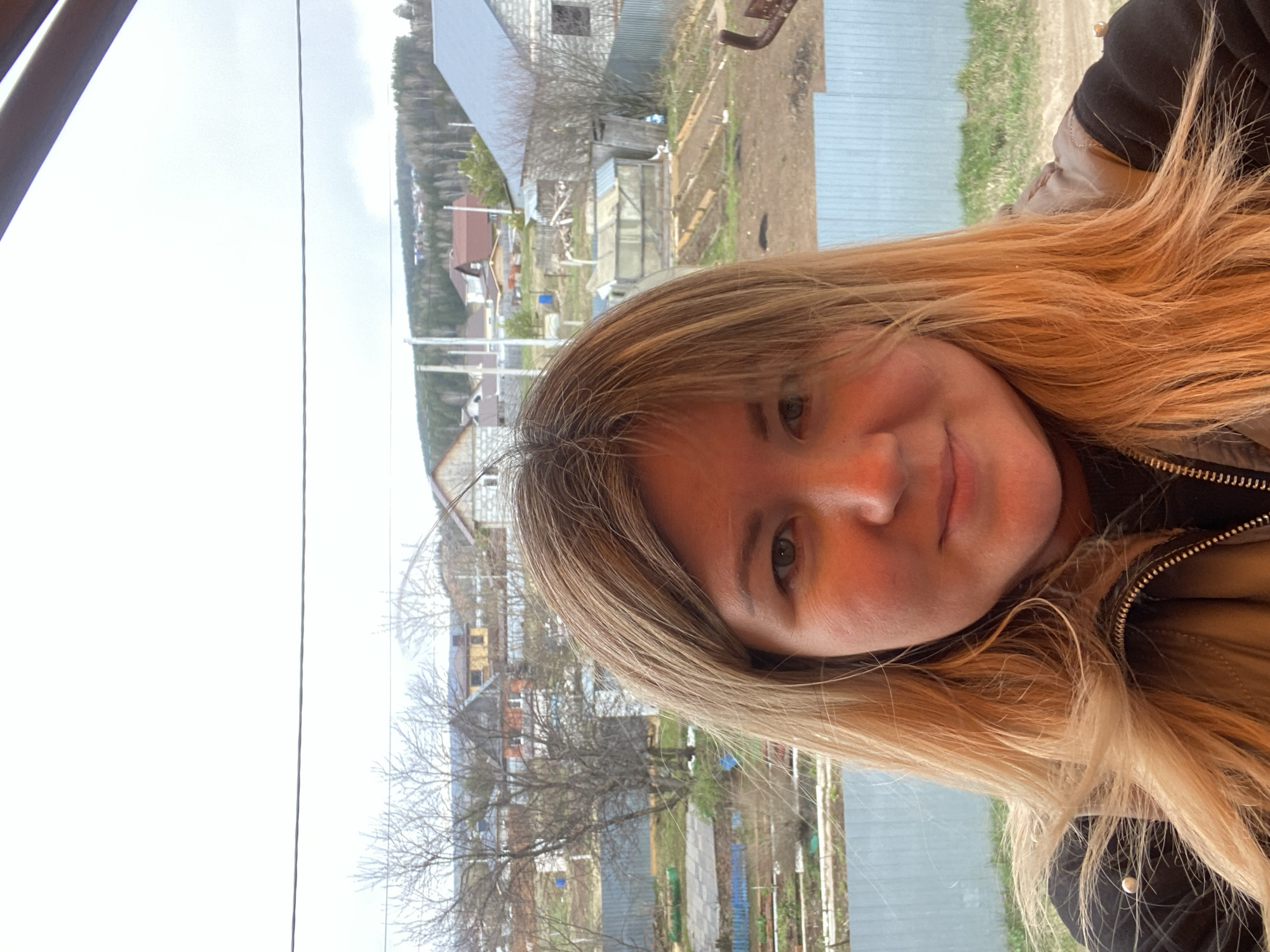 This bright, sideways-oriented image captures a woman taking a selfie with a rural landscape in the background, set in Ольховка, Russia.
The subject, a woman with long, wavy blonde hair, is positioned on the right side of the frame (when viewed upright), smiling gently towards the camera. She is wearing a dark, possibly black or brown, jacket with a prominent gold zipper. Her expression conveys contentment.
The background, visible over her left shoulder and extending across the rest of the image, depicts a village or rural area seen from an elevated vantage point, possibly through a window or from a balcony. Under an expansive, bright, and likely overcast sky, several houses and structures are visible, including one made of grey concrete blocks that appears to be under construction. Garden plots featuring greenhouses or cold frames are scattered throughout the scene. Bare trees indicate either late winter or early spring. In the far distance, hills or forested areas complete the landscape. A thin, dark wire or cable runs horizontally across the upper portion of the background.
The overall scene suggests everyday life in a Russian village during a transitional season, characterized by diffused daylight. No specific activity beyond taking the selfie is depicted, and no text is visible within the image.
