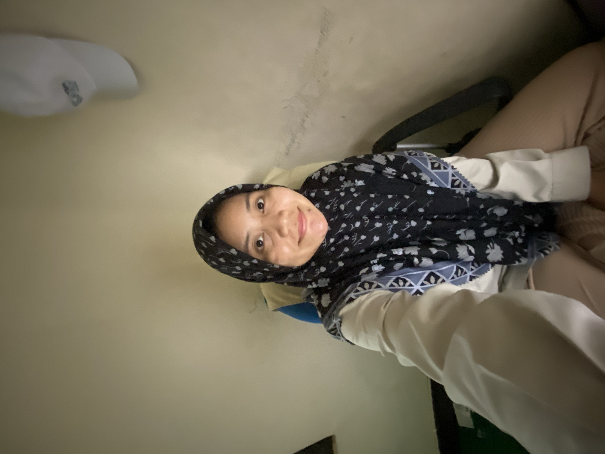 A young woman wearing a hijab and a patterned scarf smiles at the camera in an indoor setting. She is dressed in a light-colored, long-sleeved garment and tan pants. The background is a plain, neutral-colored wall with some subtle texture and discoloration. To the left of the frame, a white light fixture is partially visible on the ceiling. The lighting suggests it might be daytime or a well-lit indoor space. The overall mood appears relaxed and casual. The image is slightly tilted.