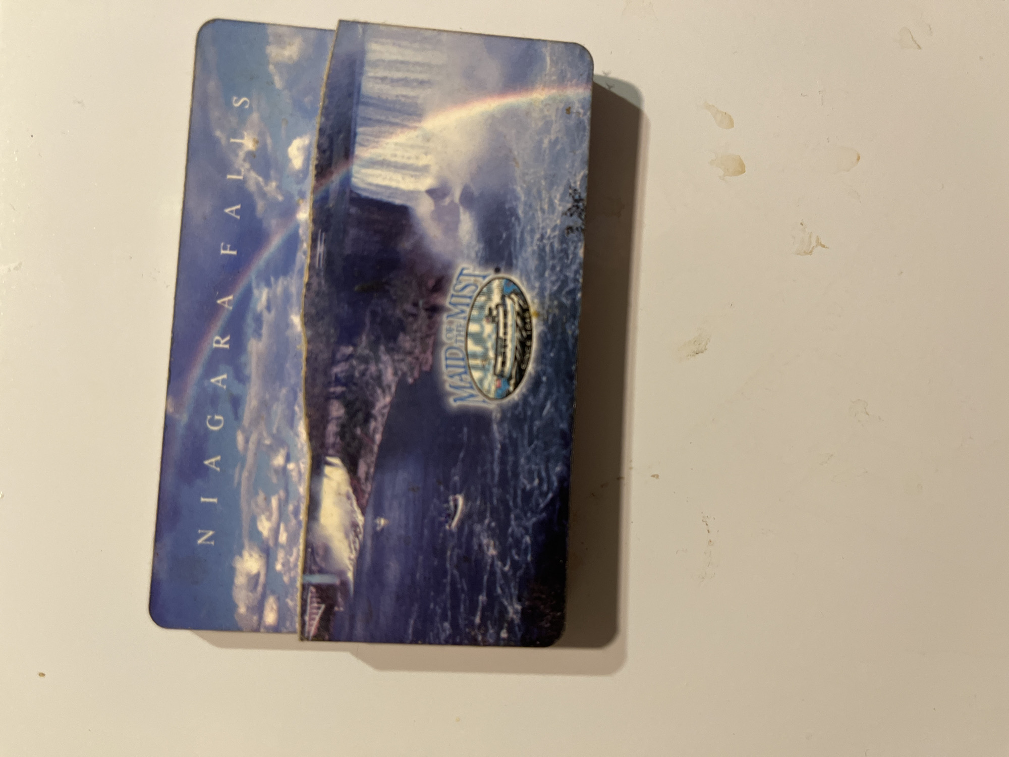A rectangular postcard or magnet, depicting Niagara Falls, is centrally positioned on a light, off-white surface. The top portion of the object displays the text "NIAGARA FALLS" in white capital letters. The image on the card features a panoramic view of the falls, with a turbulent river in the foreground, leading to the cascaded water. A prominent rainbow arcs over the mist rising from the falls, with a bridge visible to the left. In the lower-middle portion of the image, an oval logo reads "MAID of the MIST" with a depiction of a tour boat. The surrounding surface on which the object rests shows minor smudges and faint discoloration. This item is located in the city of Minsk, Belarus.