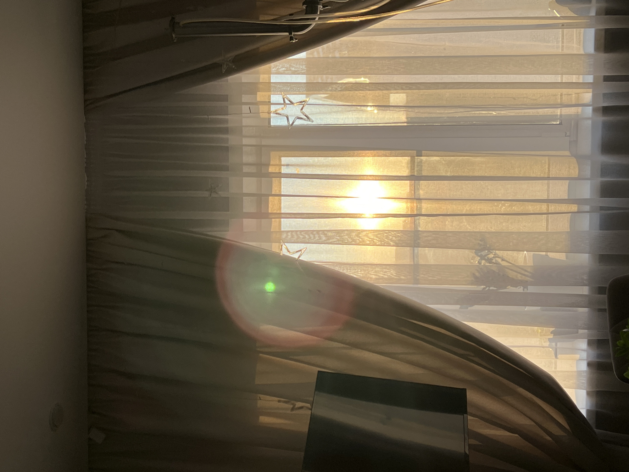 The image shows a window with sheer curtains, through which the setting sun is visible. The sun's rays create a warm, golden glow that illuminates the room. A lens flare, appearing as a circular or oval shape with concentric rings of color, is present in the upper left portion of the image, distorting the view of the curtains. A television screen is visible on the left side of the frame, partially obscured by the curtains. A potted plant is silhouetted against the light on the windowsill. Decorative star-shaped lights are hanging in front of the window. The overall scene is peaceful and serene, with the warm light creating a cozy atmosphere. The location is unknown, and there are no people or actions depicted.