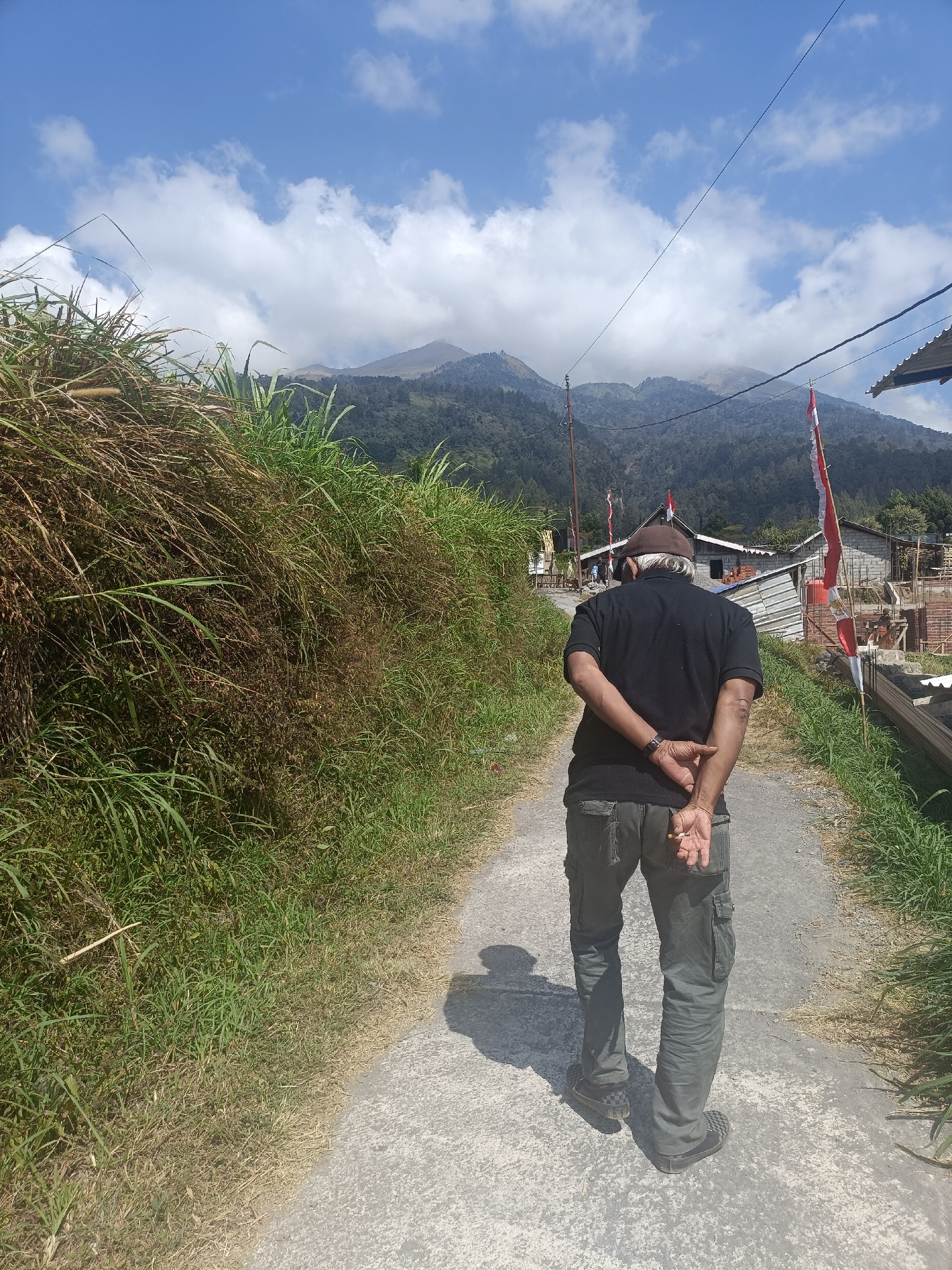 In Kopeng, Indonesia, a man walks away from the camera down a concrete path. He wears a dark t-shirt, dark cargo pants, and a brown cap. His hands are clasped behind his back, and he holds a cigarette. The path is bordered by lush green and dry grass. In the background, mountains rise under a blue sky with scattered clouds. Several buildings, some appearing under construction with visible brickwork and scaffolding, are situated along the right side of the path, adorned with Indonesian flags. Telephone wires stretch across the upper portion of the image.