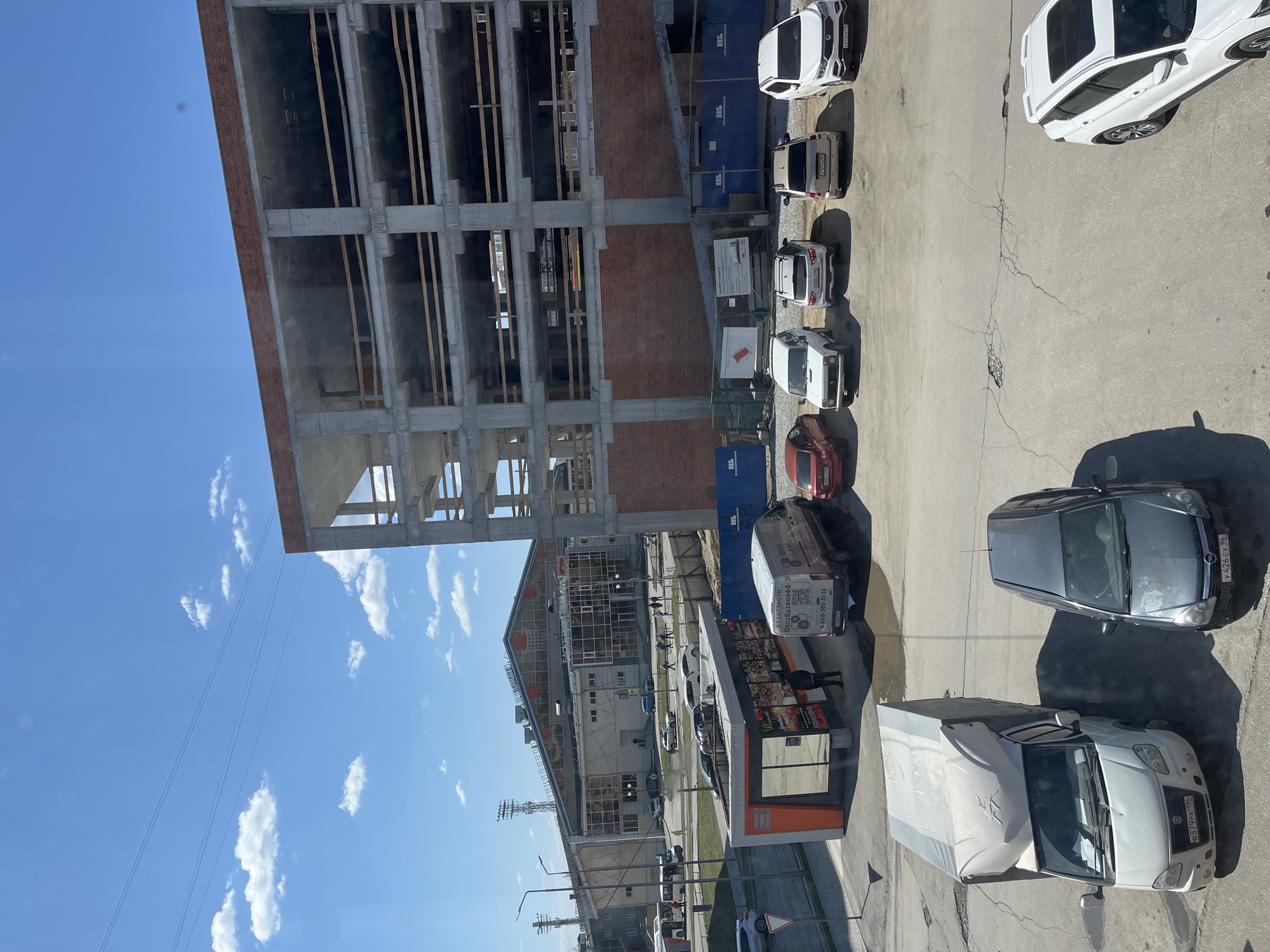 This photograph captures a scene from what appears to be Verkhnyaya Pyshma, Russia. It showcases a construction site, with an unfinished building displaying a structural framework of concrete and brick, partly visible against a blue sky dotted with clouds. To the right of the construction is a line of parked cars and a white van. Further down, there is a white truck and a silver car parked close to what seems like a roadside kiosk or stall, identifiable by the orange color on the kiosk. The kiosk is marked with some text that includes the words "БУЗАПЧАСТИ" and "GRUZRAZBOR660 GRUZ," which suggests it may be a vendor that sells replacement parts. A person can be seen standing at the kiosk's window. The image is taken from an elevated position, looking down onto the scene.