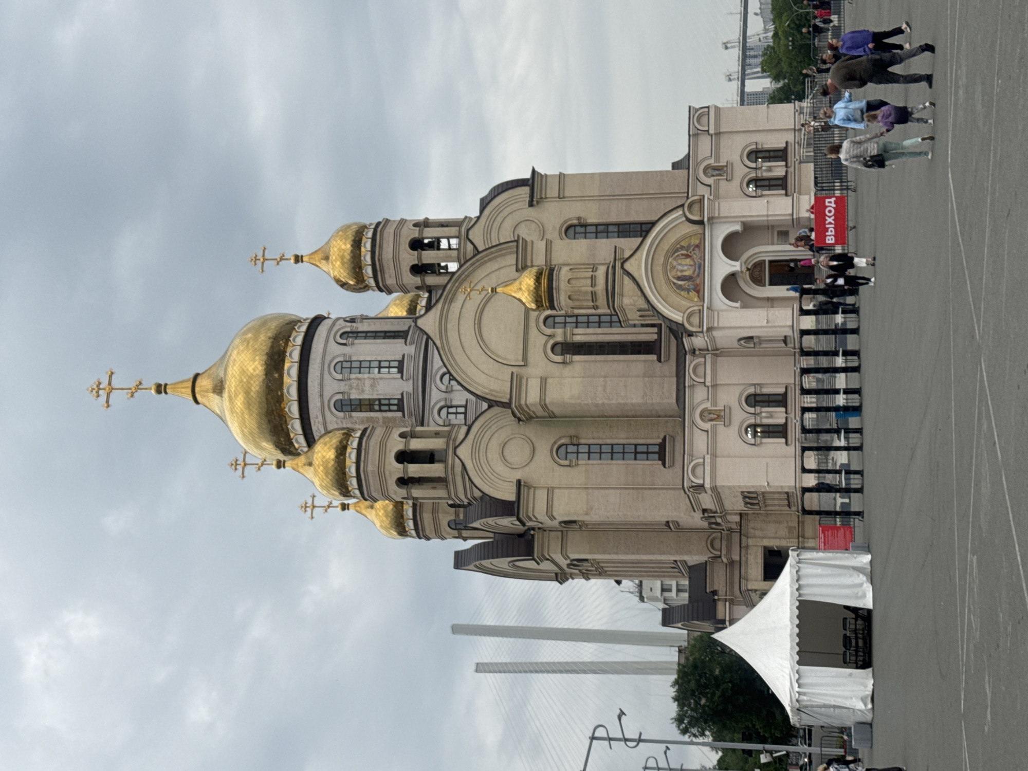 The image shows the Svetlanskaya Church of the Intercession of the Mother of God in Vladivostok, Russia. The church is a large, ornate building with multiple golden domes and crosses. In the foreground, there is a white tent, a metal detector archway, and several people walking around. In the background, a large cable-stayed bridge is visible. The sky is overcast.