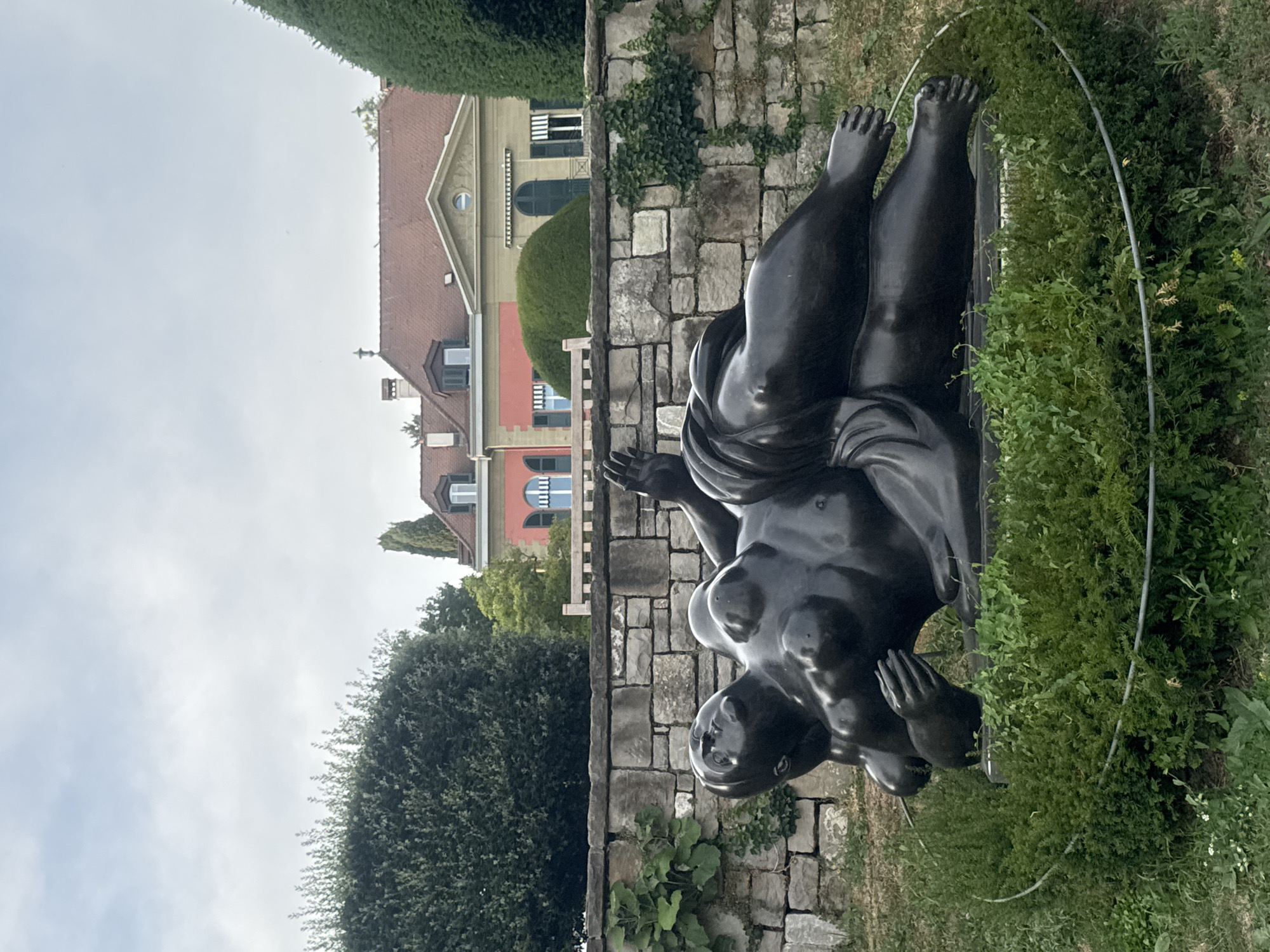 This is an outdoor daytime photograph of a large bronze sculpture of a reclining nude figure in a garden. The sculpture is positioned in the foreground, lying on its back with its legs crossed and arms raised. Behind the sculpture is a low stone wall, and beyond that, a row of buildings with red and yellow facades and pitched roofs. Green trees and manicured hedges frame the scene. The sky is overcast. This sculpture is identified as "La grande odalisque" by Fernando Botero and is located in Lausanne, Switzerland.