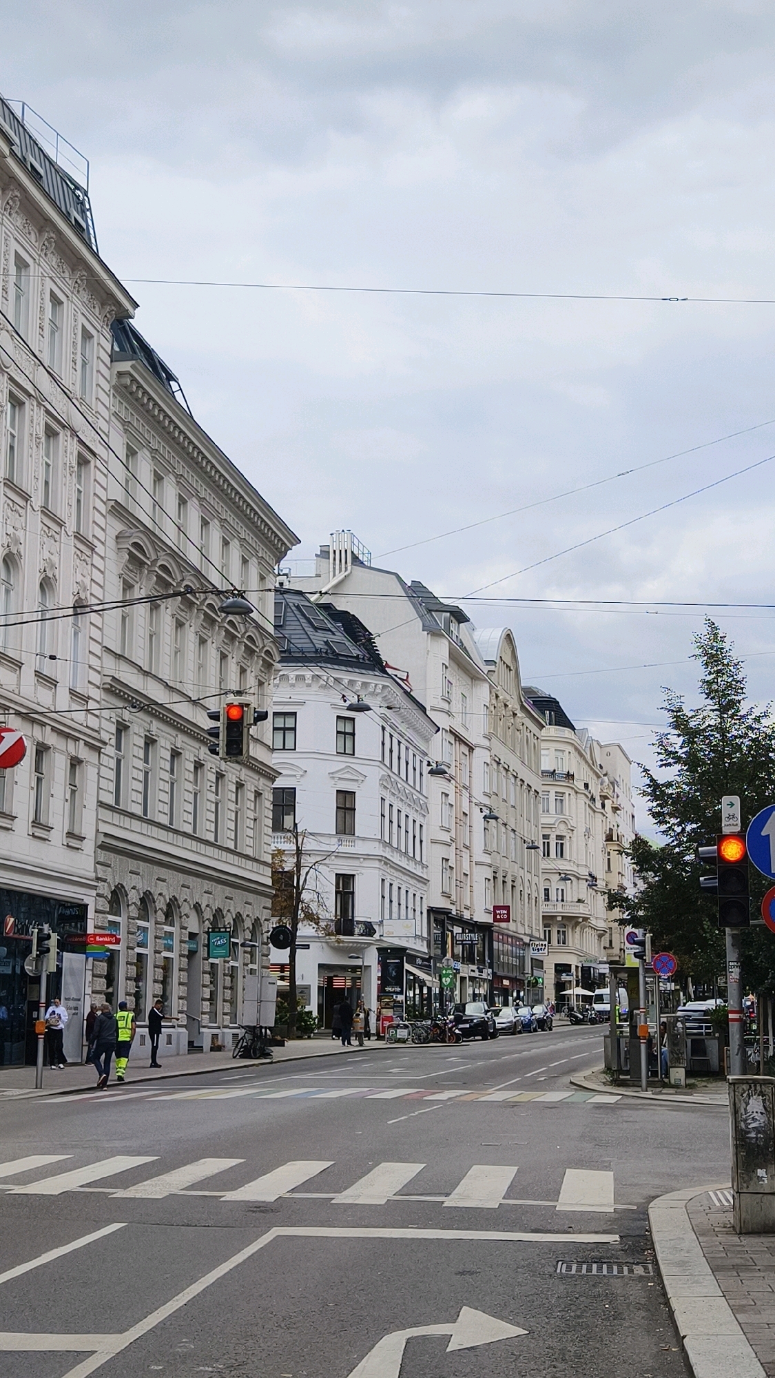 This image captures a street scene in Vienna, Austria, featuring a row of ornate, multi-story buildings lining a wide, grey asphalt road. The sky is overcast with a pale, cloudy texture. On the left side of the road, a pedestrian crossing with white stripes is visible. A traffic light on a pole displays a red signal. Several people are present on the sidewalk, including two individuals wearing high-visibility vests, one carrying a blue bag, and another walking away from the camera. A circular red sign with a white border and a red diagonal line is affixed to a pole. Further down the street, more people are visible walking and standing near shop entrances. Bicycles are parked on the sidewalk. The buildings exhibit architectural details such as arched windows, decorative facades, and pitched roofs with chimneys. Overhead, power lines crisscross the sky. A traffic light on the right side of the road also shows a red signal, accompanied by a circular blue sign with a white arrow pointing right and a bicycle symbol. A tall, dark green tree stands on the right side of the image, partially obscuring the buildings. The scene conveys a sense of urban activity on a cloudy day.