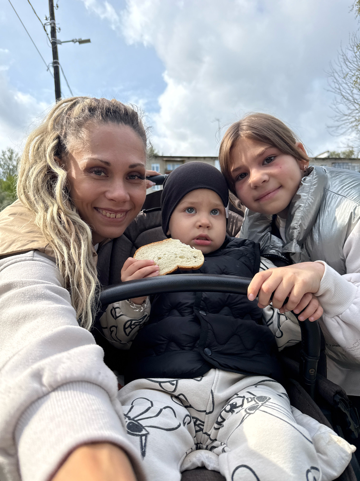 In a medium outdoor shot, a woman with blonde dreadlocks smiles at the camera, positioned on the left. Beside her and slightly in front is a young girl with brown hair, leaning in towards the camera. Between them, a baby boy, wearing a black hat and vest, is seated in a stroller, holding a slice of bread. The baby is looking upwards. The background shows a cloudy sky with some trees and buildings. The setting appears to be a street in Rusanovo, Russia.