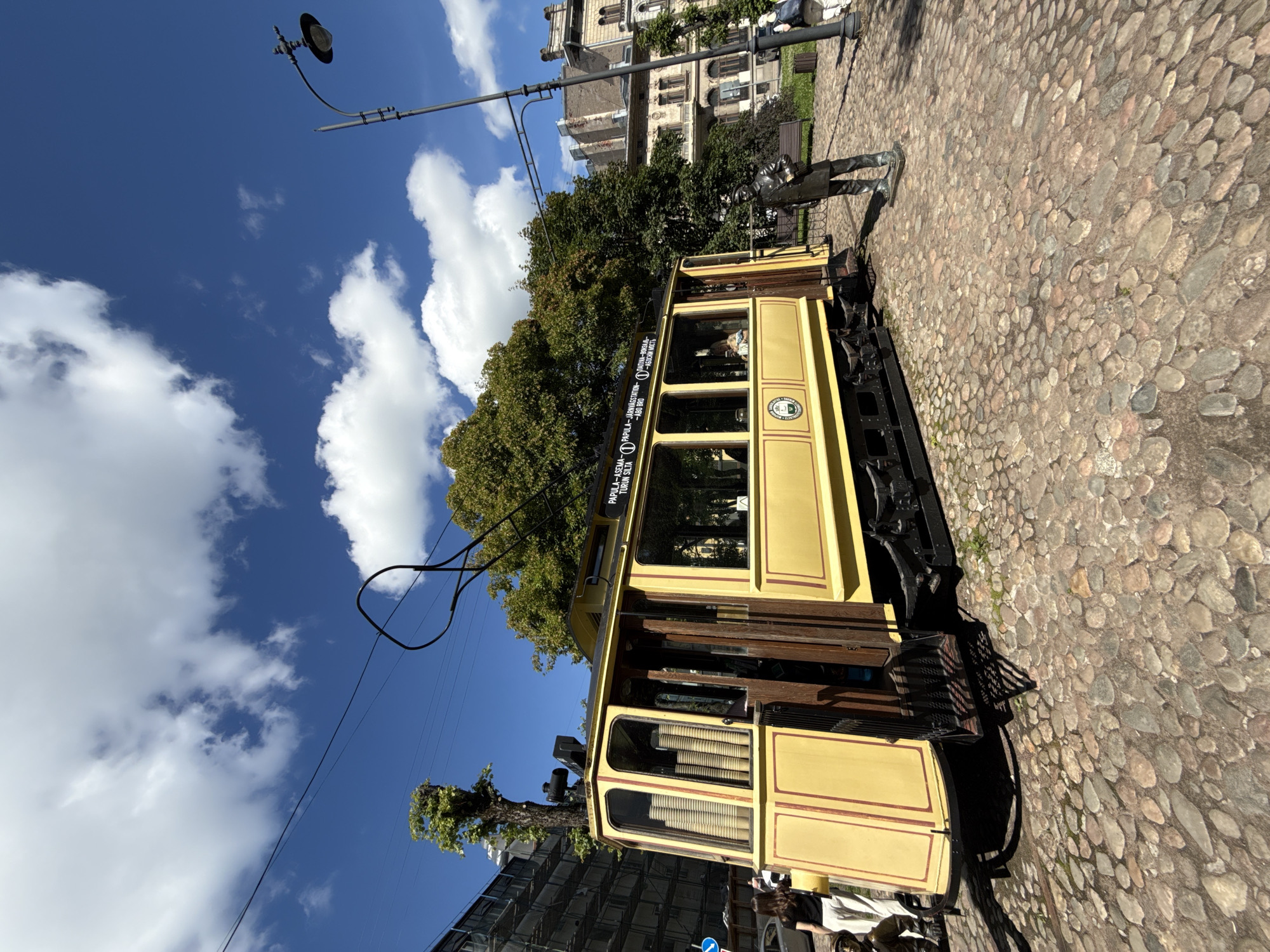A vintage yellow tram is parked on a cobblestone street in Vyborg, Russia. The tram has wooden doors and windows, and is decorated with red trim. A statue of a man in a uniform stands in front of the tram. In the background, there are trees, buildings, and a street lamp. The sky is blue with white clouds.