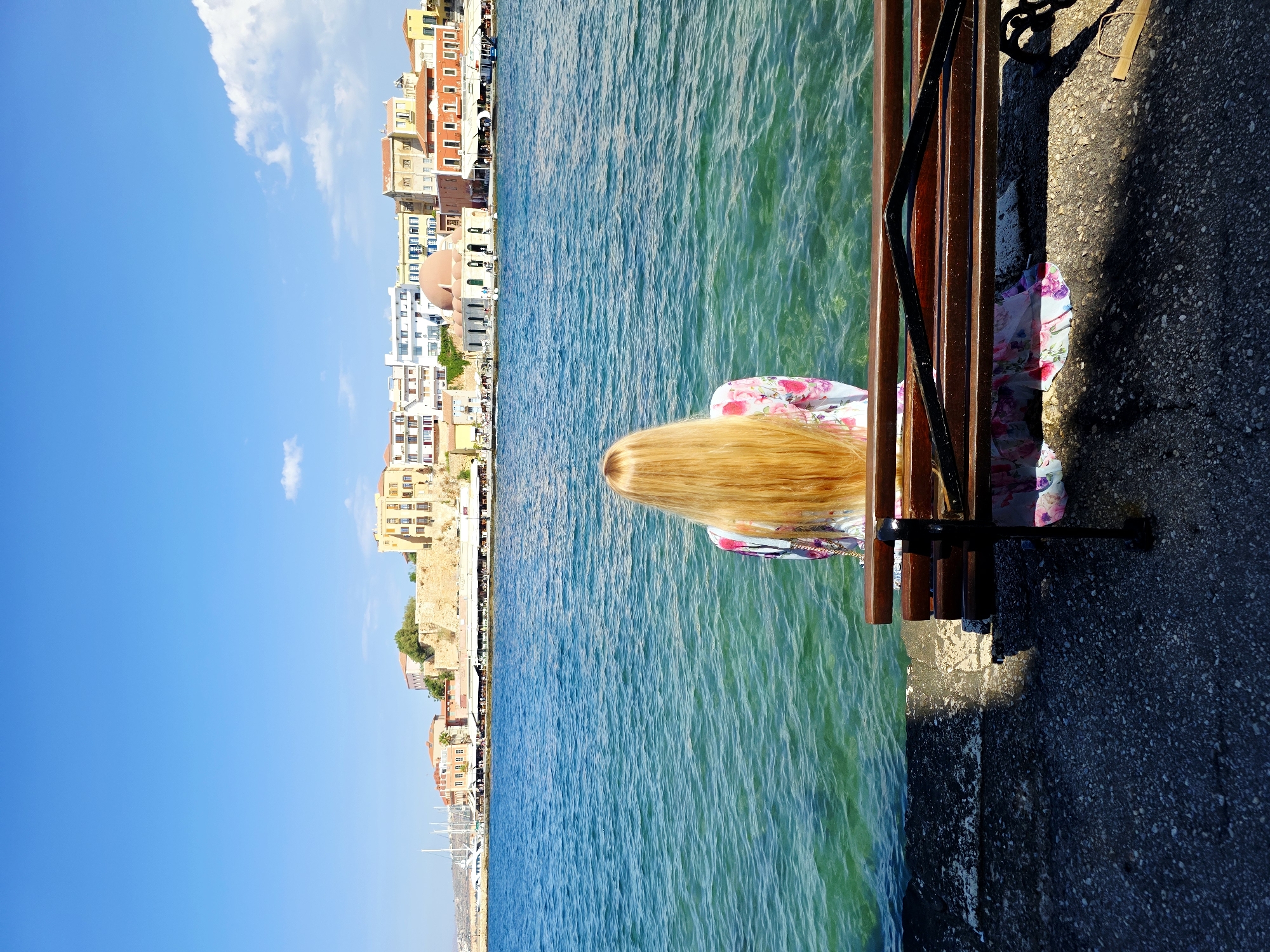 The image shows a woman with long blonde hair, viewed from behind, sitting on a wooden bench facing a body of water. She is wearing a floral dress. The water is clear and blue, with visible ripples. Beyond the water is a cityscape with several buildings, some of which are in shades of yellow, orange, and white. A prominent dome-shaped building with a pinkish-red roof is visible in the center of the cityscape. The cityscape appears to be a waterfront area with a harbor, as indicated by the presence of sailboats in the background. The foreground consists of a stone walkway and the bench. The sky is blue with a few scattered clouds. The scene is lit by bright sunlight, casting shadows on the walkway and bench. This view is characteristic of the Venetian Harbor in Chania, Crete, Greece.