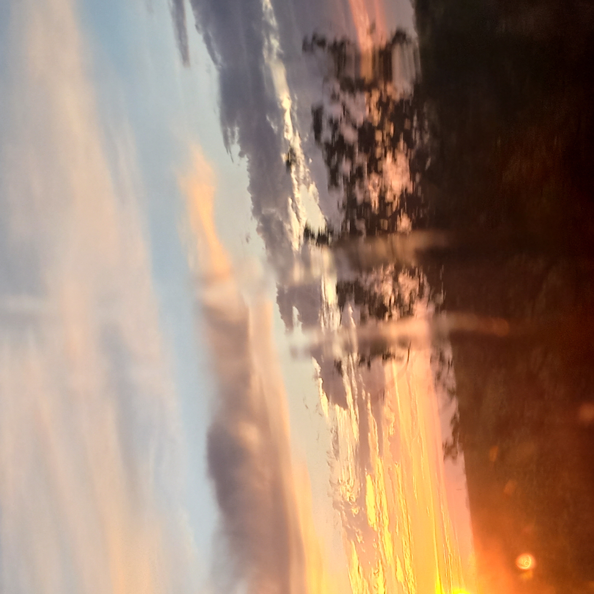 This is a blurred, horizontally-oriented image of a sunset sky with silhouetted trees in the foreground, taken from a moving vehicle, suggesting a highway or rural road setting near Kropyvnytskyi, Ukraine. The sky is a gradient of colors, starting with bright yellow and orange on the left, transitioning to pink and then a pale blue with scattered clouds. The sun itself is not clearly visible but its intense light creates a bright glow on the left side of the frame. The trees are dark and indistinct due to motion blur. There are no people or discernible structures visible in the image. The overall impression is one of fleeting natural beauty captured during transit.