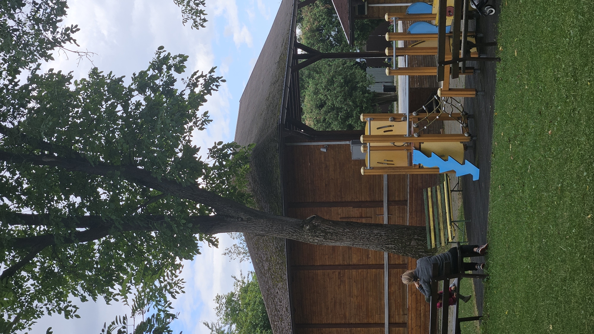 The image depicts a playground with a wooden structure and various play equipment, including slides and climbing nets. A large tree with green foliage stands in the foreground, partially obscuring a wooden building with a dark roof. Two benches are visible in the scene. On one bench, a person is seated, facing away from the camera, with another person sitting next to them. The ground is covered with grass and a dark surface. The sky is cloudy with patches of blue. This scene is located in Prešov, Slovakia, specifically at the children's playground near the park library.