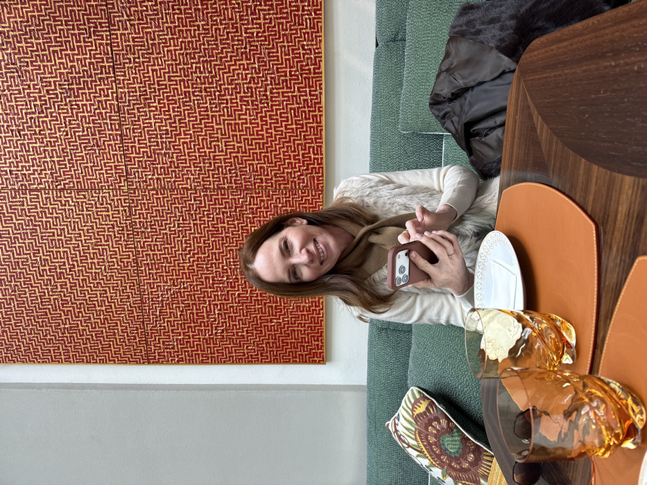 An adult female, appearing middle-aged, is seated in a sophisticated interior setting identified as the Hermès Caffè at Via Montenapoleone 12, Milan, Italy. The woman has long brown hair and a smiling expression, looking directly at the camera. She is wearing a cream-colored long-sleeved top, a light brown or beige faux fur vest, and a plain brown scarf. A ring is visible on her left ring finger. She holds a smartphone with a brown case in her left hand, with her right hand positioned as if interacting with the phone screen. In the foreground, a dark wooden table features two amber-colored, textured drinking glasses containing liquid, a white plate with an embossed rim design, a white napkin, and an orange-brown leather placemat. The woman is seated on an emerald green upholstered sofa, with a decorative patterned pillow to her left and a folded dark brown jacket to her right. Behind her, a large artwork dominates the wall, displaying a repeating gold labyrinthine pattern against a rich red background, framed by light grey walls.