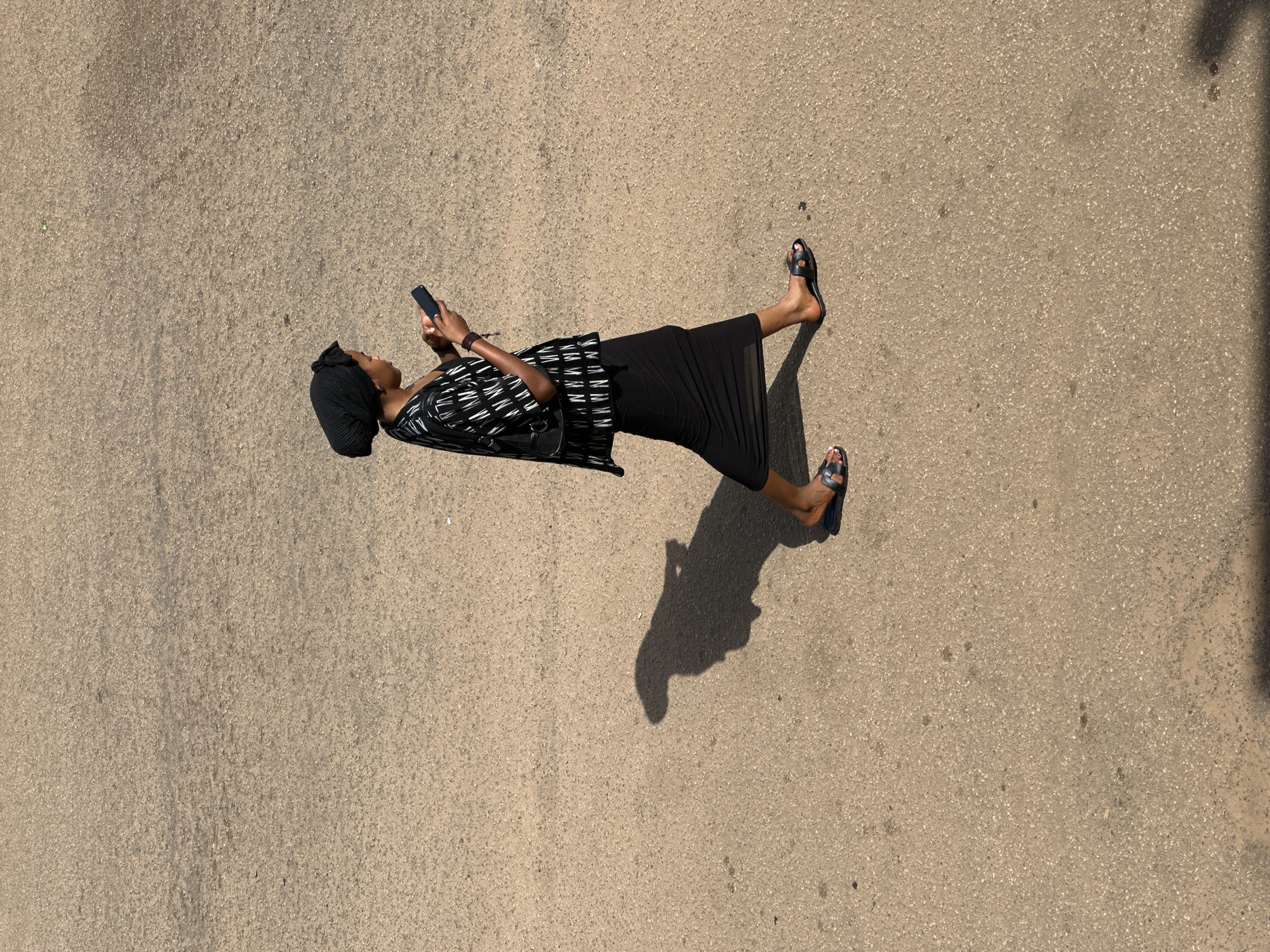 A woman wearing a black headscarf, a black and white patterned tunic, and a long black skirt walks across a paved surface, looking at her phone. She is wearing black slip-on sandals. Her shadow is cast on the ground to her left. The scene is outdoors in Bukuru, Nigeria. The paved surface appears to be asphalt or a similar material, with a rough, granular texture. The lighting suggests it is daytime.