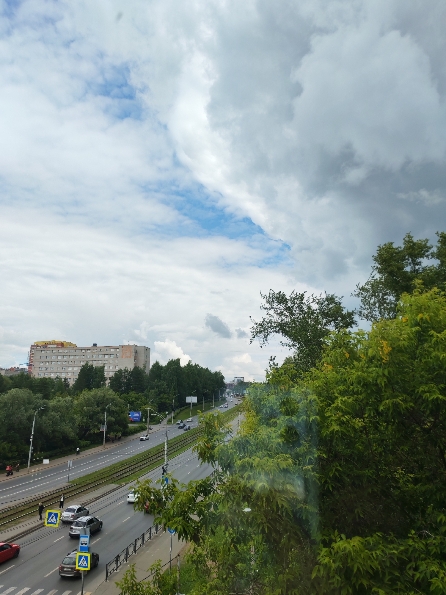 The image shows a street scene in the city of Izhevsk, Russia, with traffic, pedestrians, buildings, and lush green trees. The sky is partially cloudy, with patches of blue visible between the white and gray clouds.
In the foreground, lush green trees obscure the lower right portion of the image. A road with multiple lanes of traffic runs horizontally across the frame, with tram tracks visible between lanes. Several cars are driving on the road, and a few pedestrians are near a crosswalk. Yellow pedestrian crossing signs are visible.
In the middle ground, a long, multi-story building, likely an apartment complex or dormitory, is situated behind a line of trees. The building is light-colored with rows of windows and a yellow-colored upper section. Further down the road, more buildings and trees are visible.
The background features a distant cityscape under a dramatic sky. The overall impression is a busy urban environment integrated with natural elements.