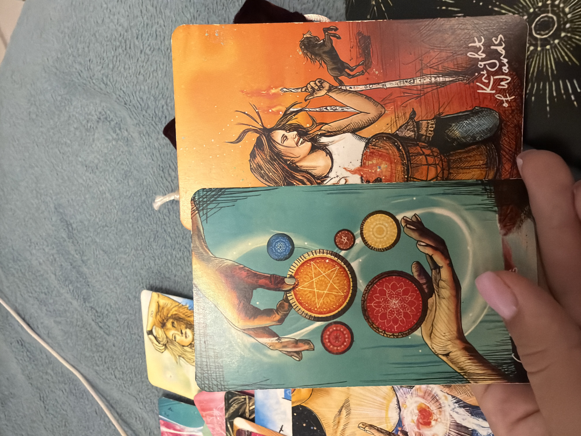The image displays a close-up view of tarot cards laid out on a soft, blue fabric surface, possibly a blanket or towel. A white cord runs diagonally across the upper left portion of the frame.
In the foreground, a hand with light pink nail polish is holding a tarot card. This card features a turquoise background with two hands and several circular symbols, some resembling coins with intricate patterns. The hand holding the card appears to be part of the illustration on the card itself.
To the right of this card, another tarot card is visible. This card has an orange background and depicts a woman with dark hair and a smiling expression, holding a drum and a wand. A dark silhouette of a horse is also present in the background, and the text "Knight of Wands" is written in the bottom right corner. Other tarot cards are partially visible in the background, suggesting a tarot reading.
The setting does not provide any specific information or clues about a location in Sharm El Sheikh, Egypt. The scene is focused entirely on the tarot cards and the hand interacting with them.