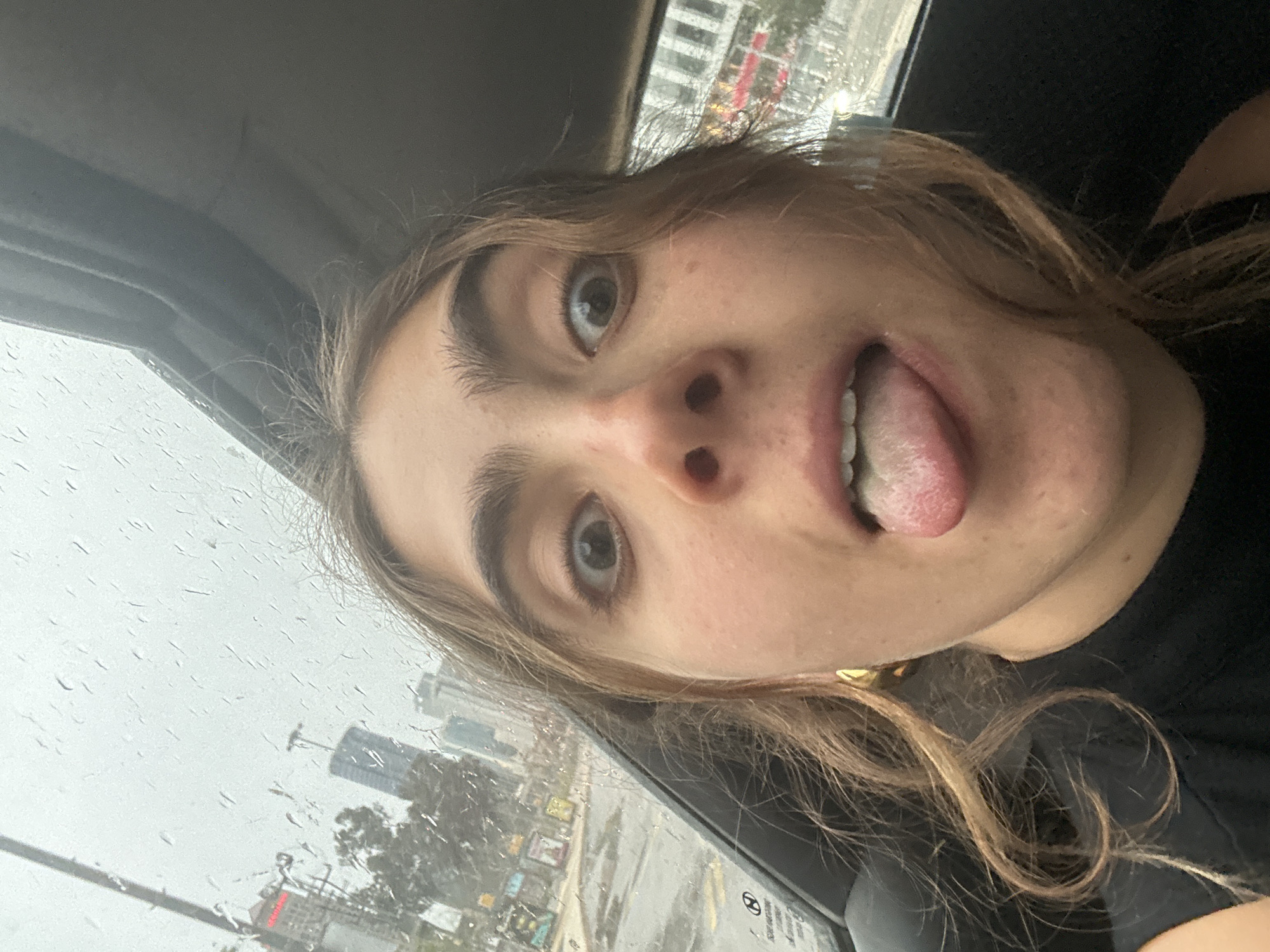 A woman is taking a selfie from inside a car. She is fair-skinned with light brown hair, protruding her tongue and looking up towards the camera. She is wearing a black shirt. The background reveals a rainy day in what appears to be Tel Aviv, Israel. High-rise buildings are visible outside the rain-streaked window, suggesting a modern urban landscape.
