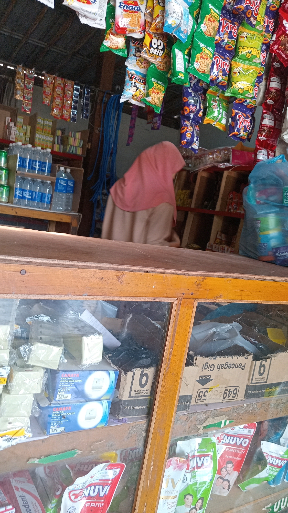 The image captures a view inside a small convenience store, likely in Kedungwaru, Indonesia. The foreground features a wooden counter with a glass display case, revealing various packaged goods including what appear to be soap bars and laundry detergent. Behind the counter, shelves are stocked with a wide array of snacks in colorful packaging, many hanging from the ceiling. Prominently displayed are bottles of water on a shelf. A person wearing a pink hijab is visible behind the counter, suggesting they are likely the shopkeeper or an employee. The overall impression is one of a typical local shop, well-stocked with everyday items for sale.