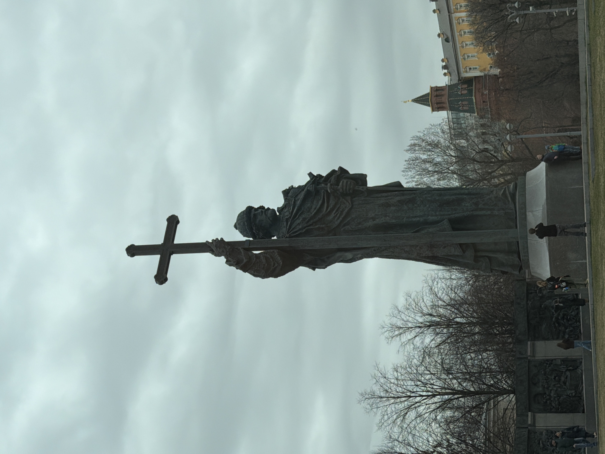 The image displays the monumental bronze statue of Prince Vladimir the Great, located on Borovitskaya Square in Moscow, Russia. The statue depicts a male figure, clad in robes and a hat, holding a large Christian cross aloft in his right hand, with a smaller object held in his left hand against his chest. It stands on a multi-tiered pedestal adorned with bas-reliefs. The sky is overcast with heavy cloud cover. Approximately ten individuals are present around the base of the monument on a grassy area; some are observing or photographing the statue. In the background to the right, the red brick Borovitskaya Tower of the Moscow Kremlin, featuring a green and gold tented roof, is visible, alongside a pale yellow building. Bare deciduous trees surround the monument and buildings.