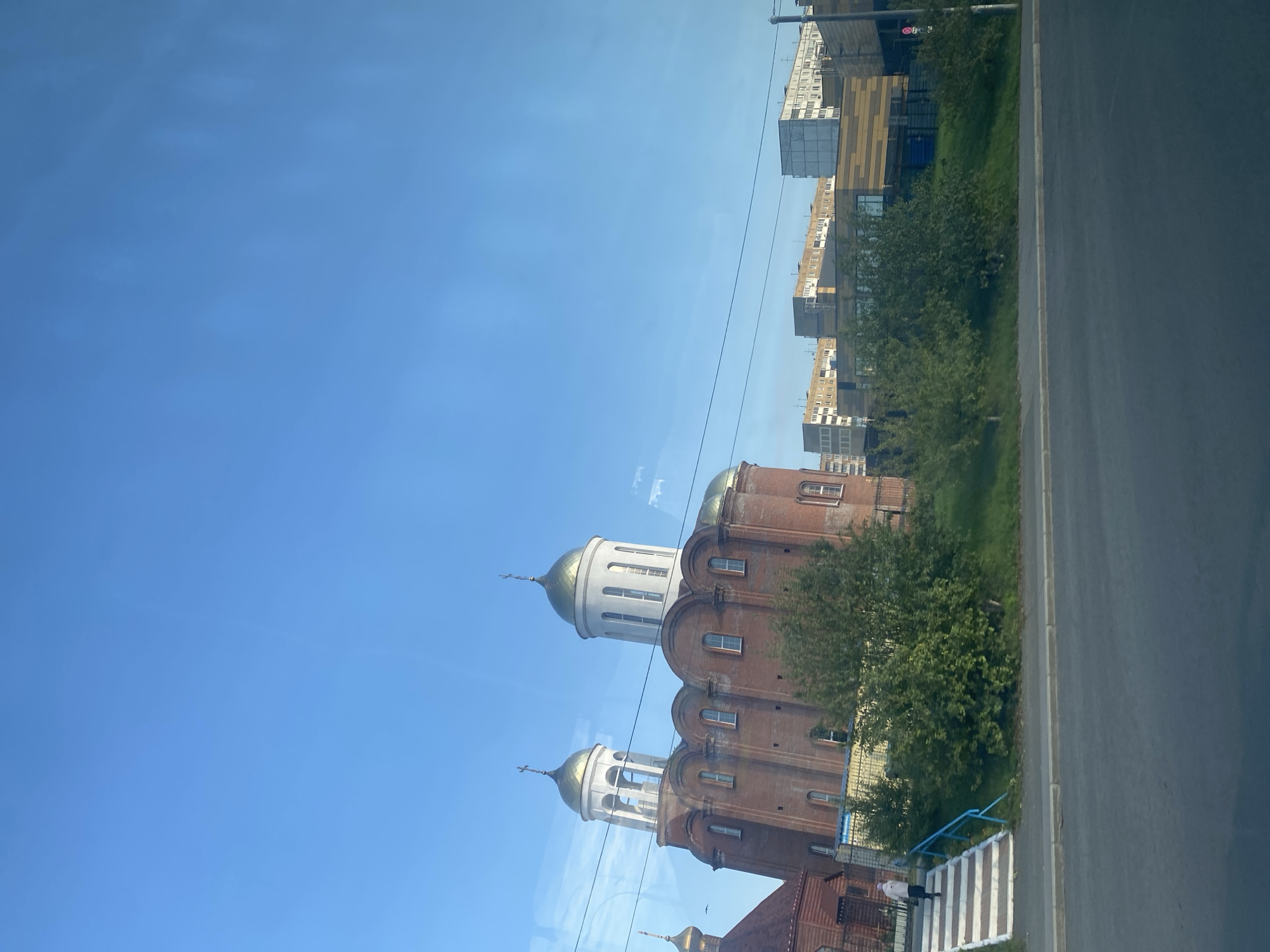 In the city of Kemerovo, Russia, a medium-angle shot captures a brick church with golden domes against a clear blue sky. To the left of the church, a smaller wooden structure with a golden dome is visible. A set of white and orange striped stairs leads up a grassy embankment towards the church. A lone figure, partially obscured by distance and angle, is ascending these stairs. In the background, beyond the church and embankment, several modern, multi-story buildings stand, suggesting an urban setting. The foreground displays a paved road and a sidewalk, implying the shot was taken from a moving vehicle or the side of the road.