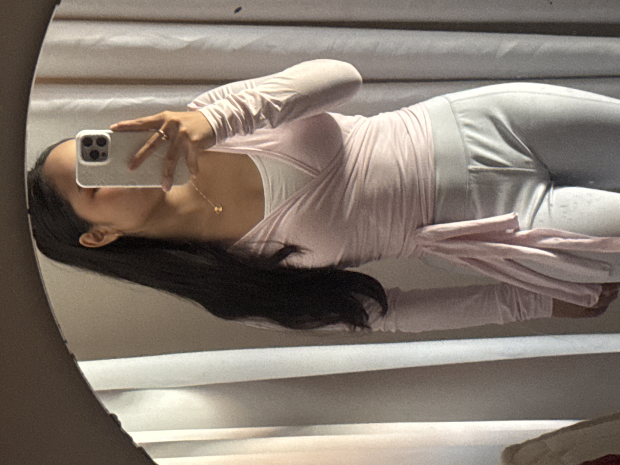 A person takes a mirror selfie. The person has long, dark hair, light skin, and is wearing a pink wrap top over a white tank top and light gray leggings. They are holding a smartphone with a light-colored case. The background appears to be a window with blinds. The image appears to be taken indoors. The image is taken in a round mirror. No specific details indicate the location is Davao City, Philippines.