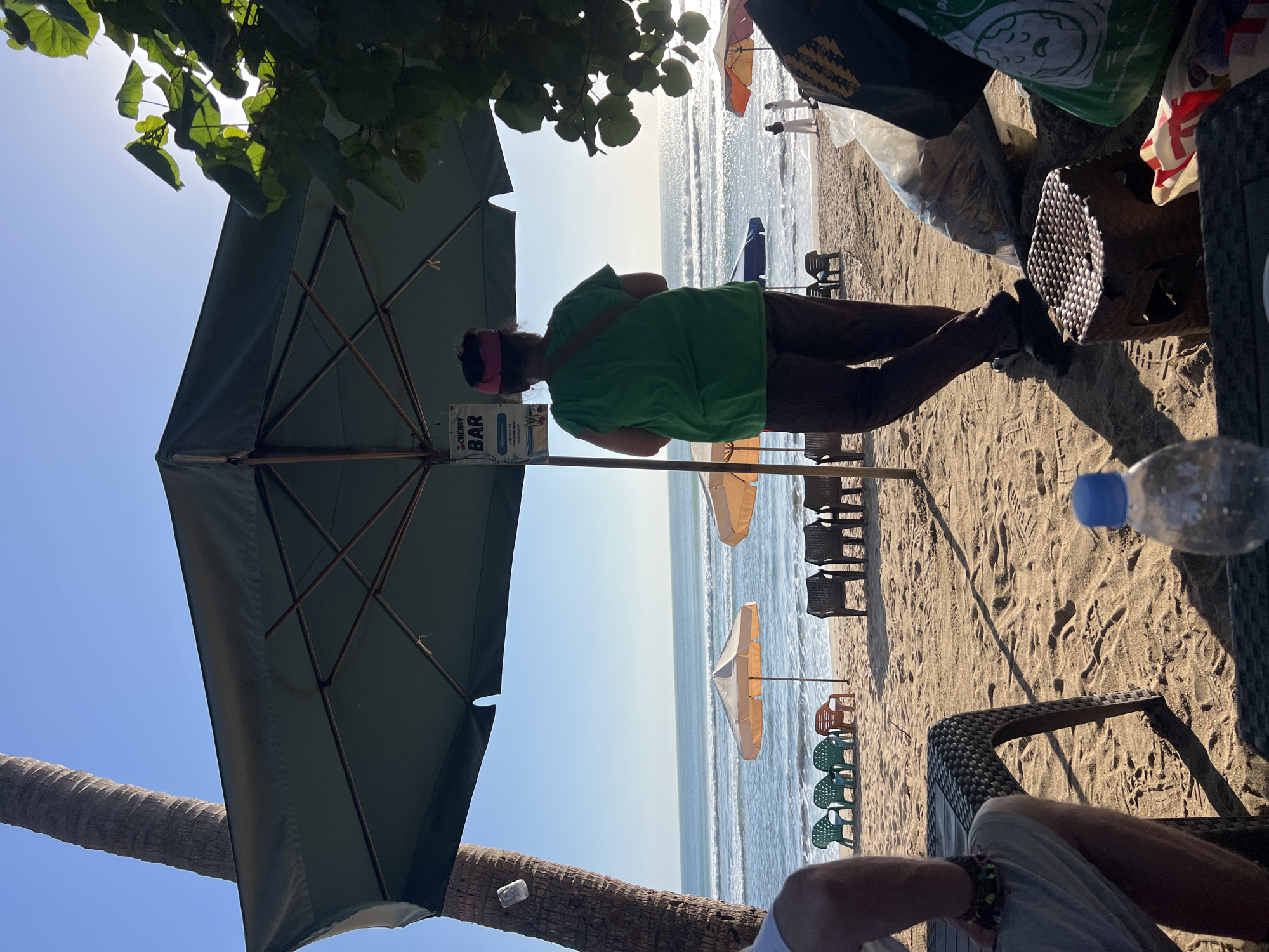 A full shot shows a beach scene in Kuta, Indonesia, with the ocean in the background. In the foreground, a person in a green shirt and brown pants stands under a large, dark green umbrella. They are looking out towards the ocean. A sign for "CHERRY BAR" hangs from the umbrella. To the left, a palm tree trunk is visible. On the beach, there are several empty chairs and umbrellas. A plastic bottle of water and some bags are on the sand. The sun is shining brightly, and the ocean has small waves.