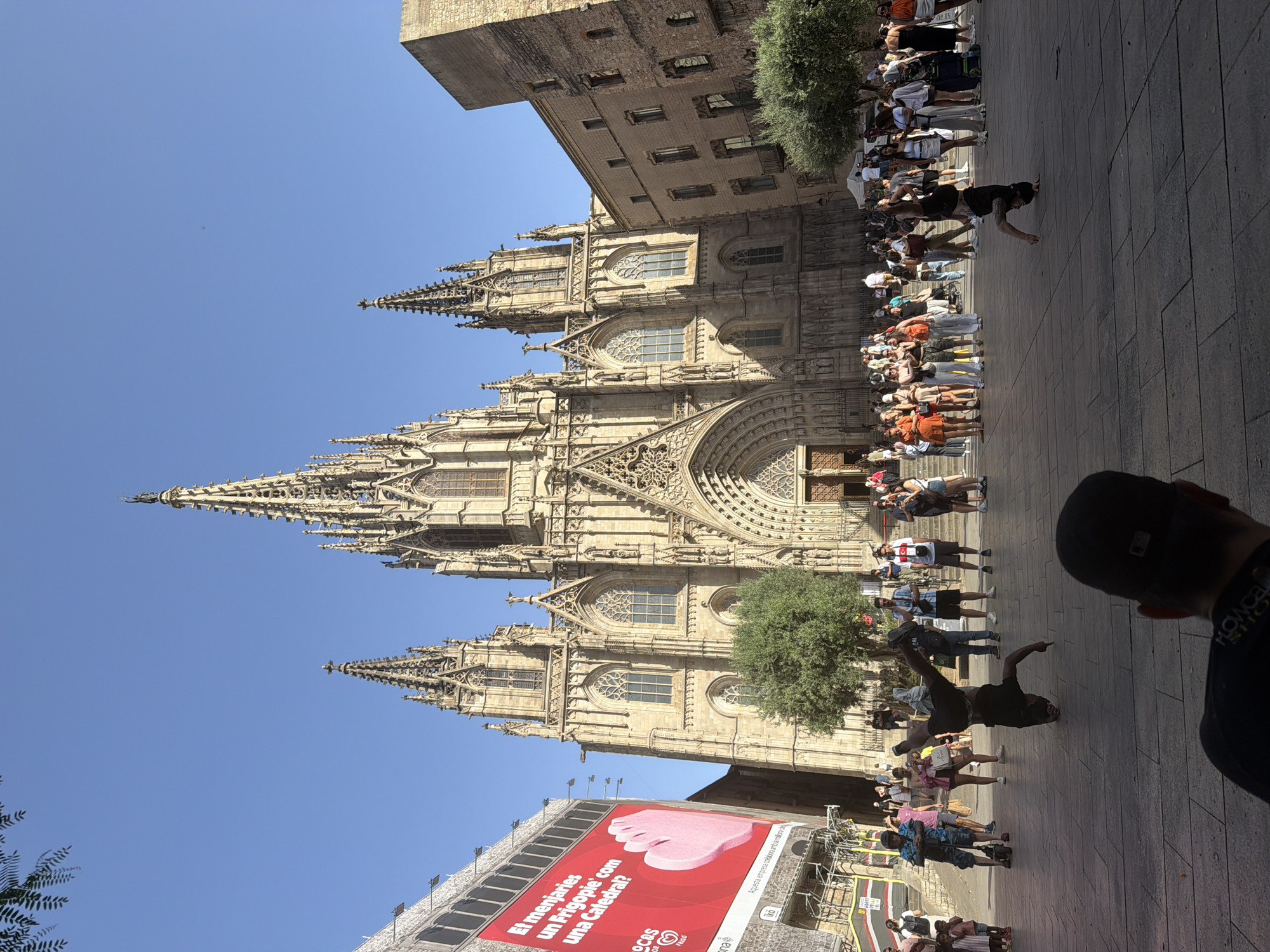 The image depicts a bright, sunny day in Barcelona, Spain, in front of the Barcelona Cathedral. The Gothic cathedral, with its intricate spires and stonework, dominates the background. To the left, a large red billboard with text in Catalan and an image of a pink hand is displayed on a scaffolding structure, suggesting ongoing construction or advertising. In the foreground, the paved plaza is populated with a diverse crowd of tourists and locals. Two individuals are performing breakdancing moves, one in a handstand with legs extended, and another in a similar pose further to the right. The atmosphere is lively, with people casually strolling, observing the performers, and taking in the architectural grandeur of the cathedral. Trees are sparsely planted in the plaza, providing some shade. The clear blue sky indicates fair weather.