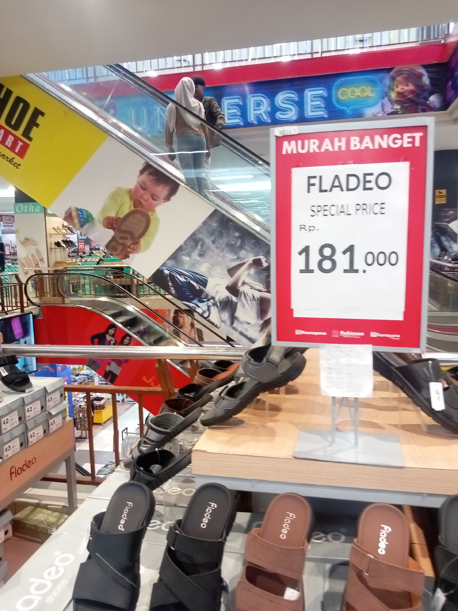 The image depicts an interior view of a shoe store, likely within a shopping mall in Batam, Indonesia, given the visible signage in Indonesian. In the foreground, a display of various sandals is prominently featured on shelving units. These include black sandals with a modern design and brown sandals with a more casual look, all bearing the "Fladeo" brand name. To the left, stacks of shoe boxes labeled with sizes like "42" and "43" are visible, also indicating the "Fladeo" brand.
In the mid-ground, an escalator ascends, with people walking on it. A large advertisement banner, primarily yellow and white, is attached to the side of the escalator, featuring a graphic of a baby holding a shoe. Above the escalator, a prominent red sign with white text declares "MURAH BANGET" (Very Cheap) and advertises "FLADEO" at a "SPECIAL PRICE" of "Rp. 181,000". Other signs, including one with "COOL", are visible in the background. The overall environment suggests a retail space, likely part of a department store or a dedicated shoe outlet, characterized by bright lighting and a focus on merchandise display.