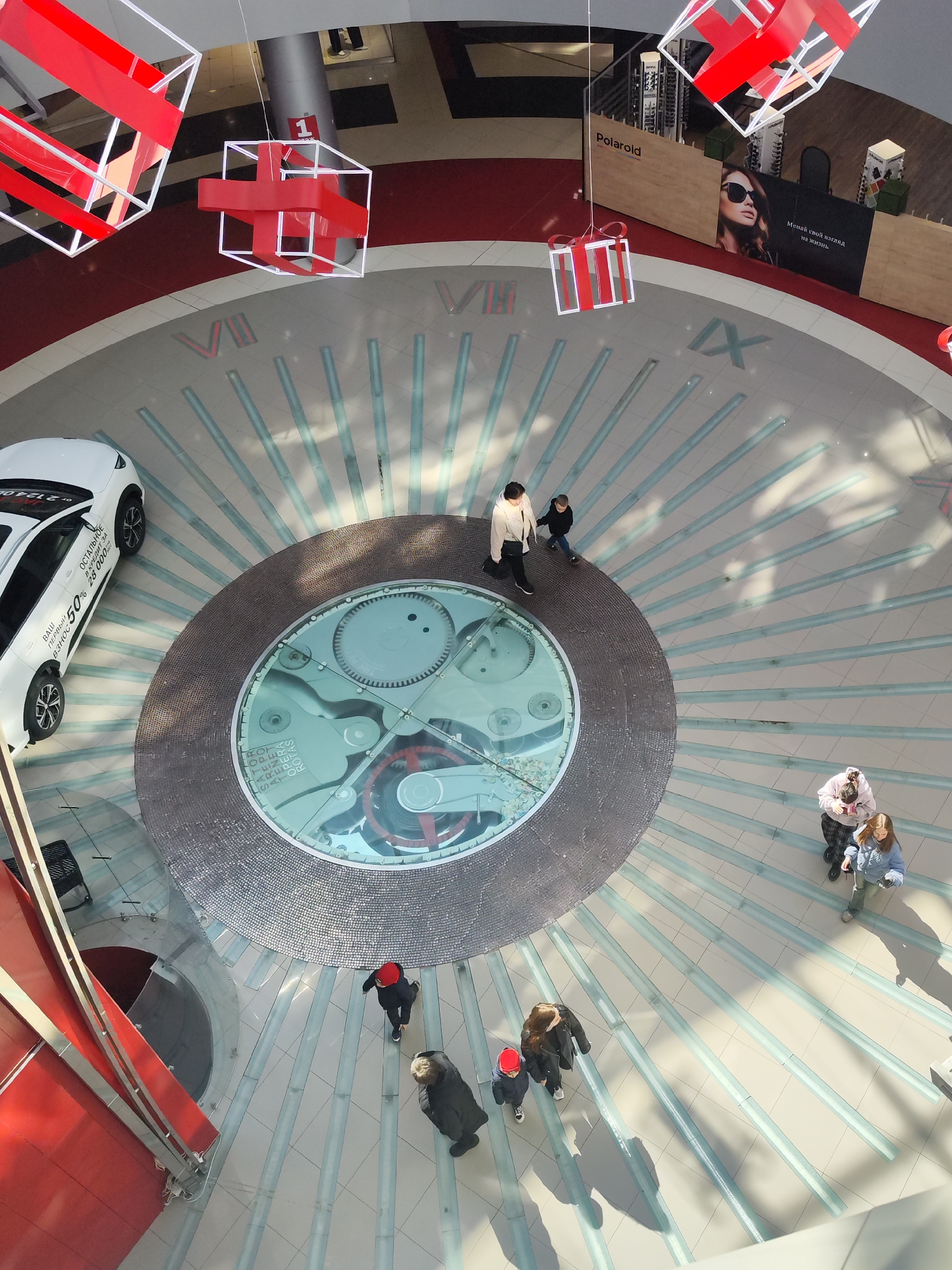 Captured from an elevated perspective, the image showcases the interior of a shopping center located in Izhevsk, Russia. The focal point is a large, decorative clock on the floor, surrounded by people walking about. The design incorporates Roman numerals to mark the hours, and there is a glass section revealing clockwork mechanisms beneath. Above, suspended from the ceiling, are decorative light fixtures shaped like red and white gift boxes, contributing to a festive atmosphere. A white car is parked nearby, advertised with a 50% off promotion in Russian. Banners for Polaroid are visible in the background.