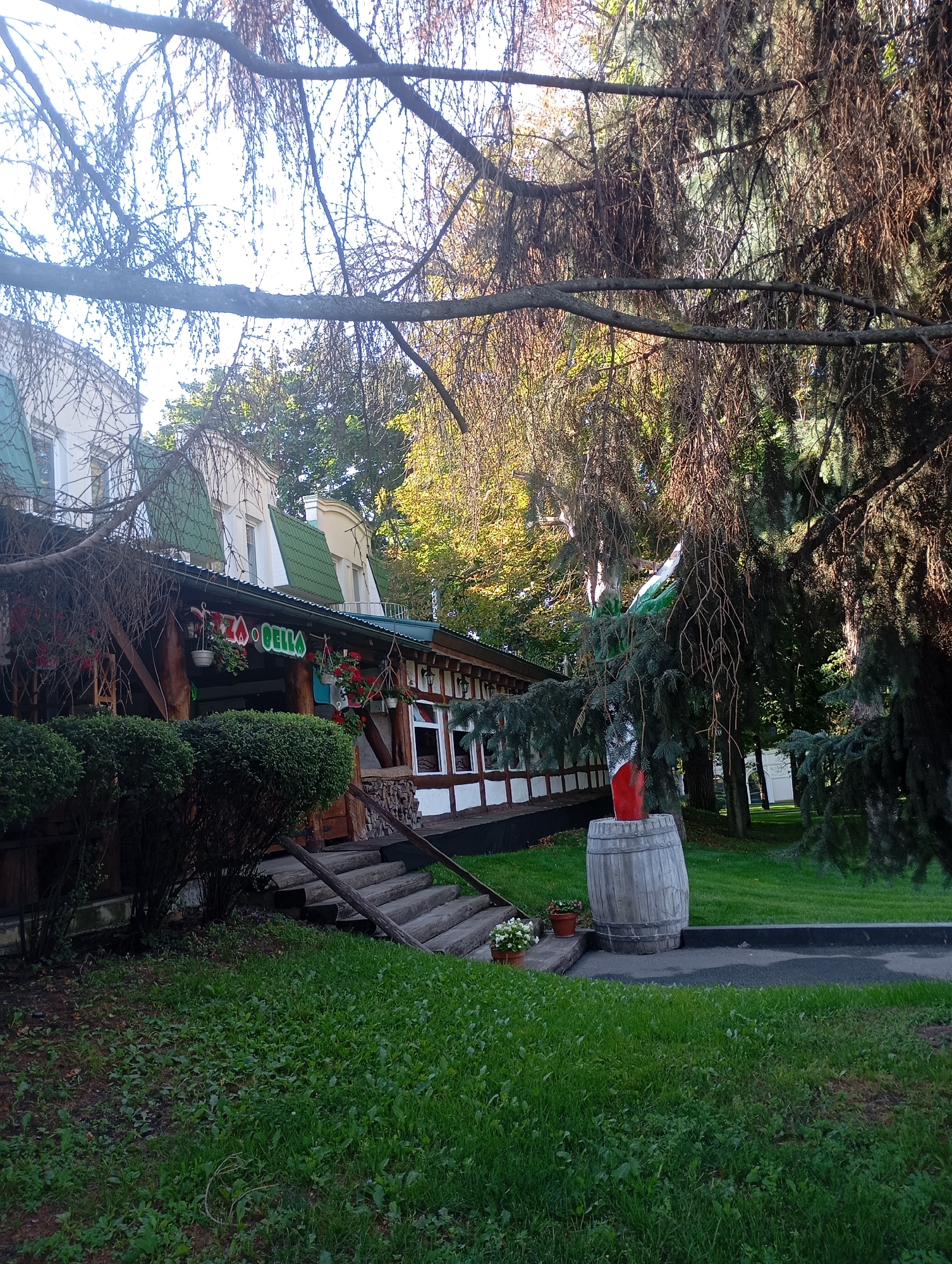 The image captures the exterior of a restaurant named "PIZZA BELLA" in Kharkiv, Ukraine, on a sunny day. The building features a green tiled roof and white and brown wooden walls. The entrance is adorned with hanging flower baskets and a sign that reads "PIZZA BELLA" in colorful letters. In front of the building, there are well-maintained green bushes and a wooden staircase leading up to the entrance. To the right of the building, a large wooden barrel sits on the grass, with a red object peeking out from its top. The surrounding area is grassy, with mature trees providing shade. The image appears to be taken from a slightly low angle, looking up towards the restaurant and trees.