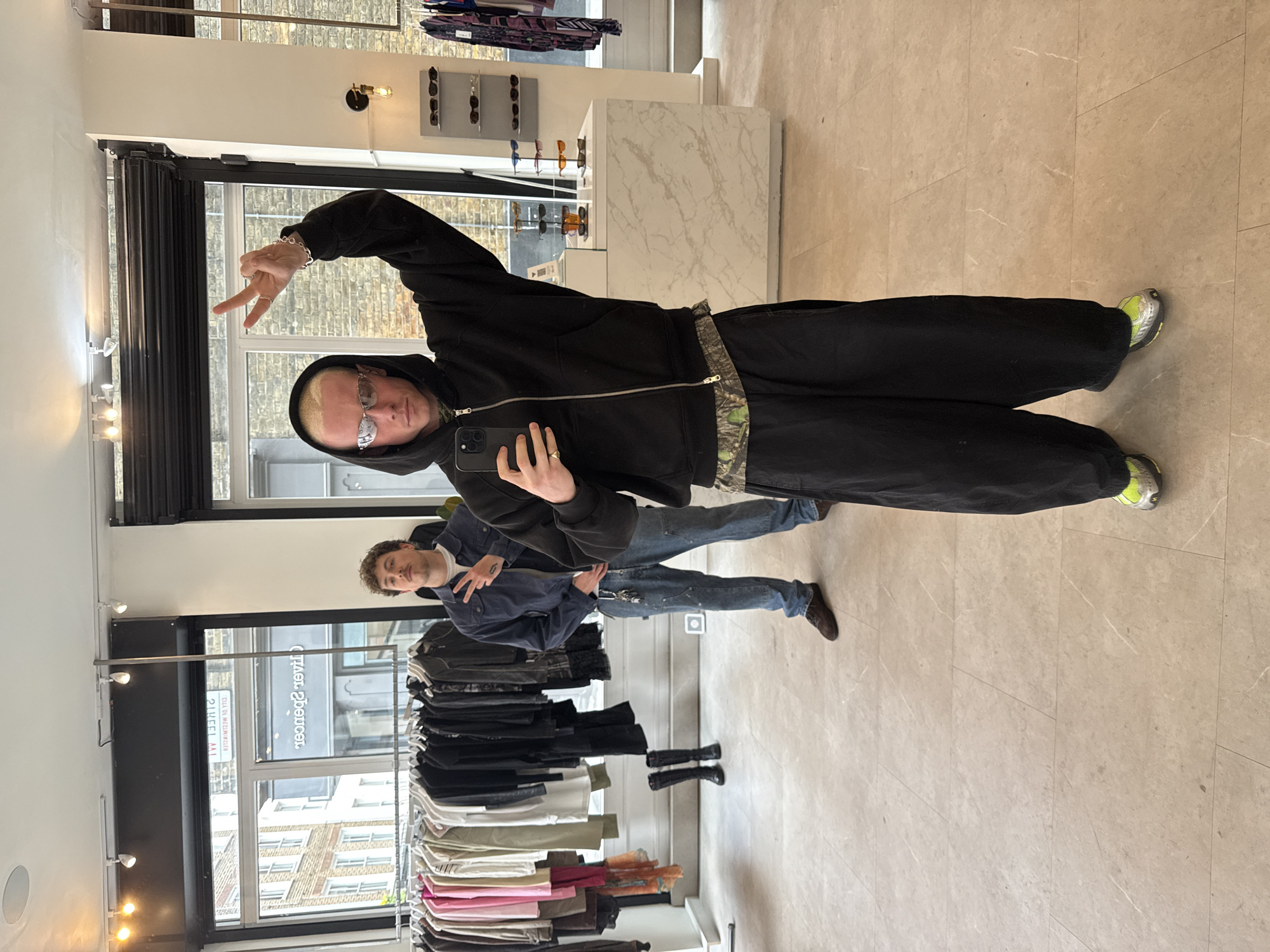 This image captures a selfie taken inside a retail store, likely a clothing or eyewear boutique. The main subject, a young man with blonde hair and glasses, is positioned on the left side of the frame. He wears a black hooded jacket with a camouflage pattern visible underneath, dark trousers, and neon yellow and grey sneakers. He holds a smartphone (appearing to be an iPhone) in his right hand and makes a peace sign with his left, showcasing a silver chain bracelet and rings.
Reflected in a large mirror behind him, on the right, is a second young man with curly dark hair, dressed in a dark blue jacket, jeans, and brown shoes. He is also making a peace sign. The store's interior features a light-colored tiled floor, white walls, and various displays, including sunglasses on shelves, a white marble-patterned counter, and racks filled with diverse garments. Natural light illuminates the scene through large windows that reflect the brick facades of buildings outside, indicating it is daytime. The overall ambiance is casual. Visible text on the window reflection reads "Oliver Spencer" and "212 BERWICK ST", confirming the store's identity and its location on Berwick Street, London.