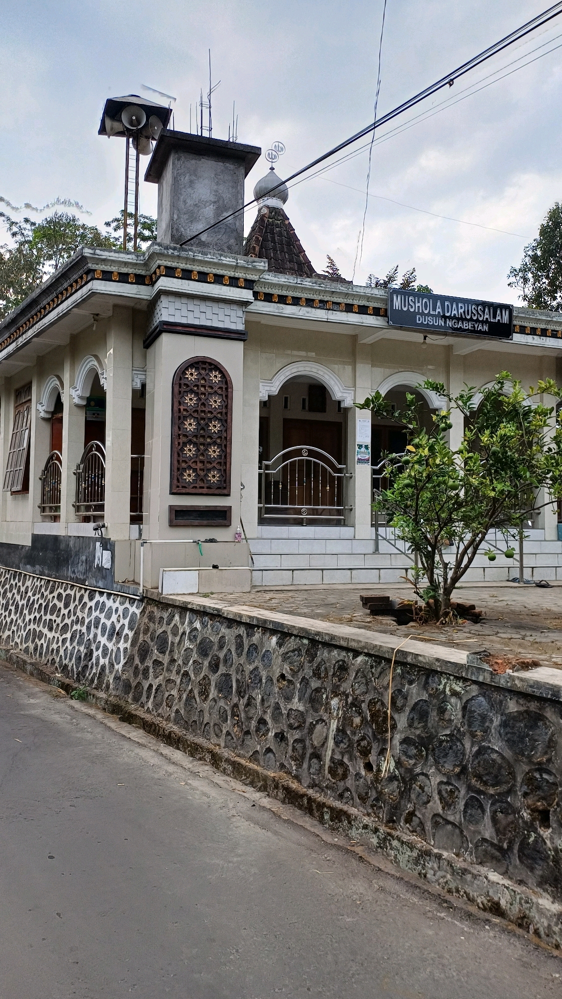 An exterior, eye-level, full shot captures a modest mosque on an overcast day in Tengahan, Indonesia. The mosque, identified by a sign as "MUSHOLA DARUSSALAM DUSUN NGABEYAN," is light beige with decorative golden trim along the roofline. It features arched doorways and windows, some with intricate wooden latticework. A small dome with Islamic calligraphy crowns the structure. Loudspeaker horns are mounted on a tall metal pole near the roof's edge. A low, rough-hewn stone wall borders the property, separating it from a paved road. A small tree with green fruit grows in front of the mosque. The sky is a muted grey, indicating cloudy weather. There are no people visible in the frame.
