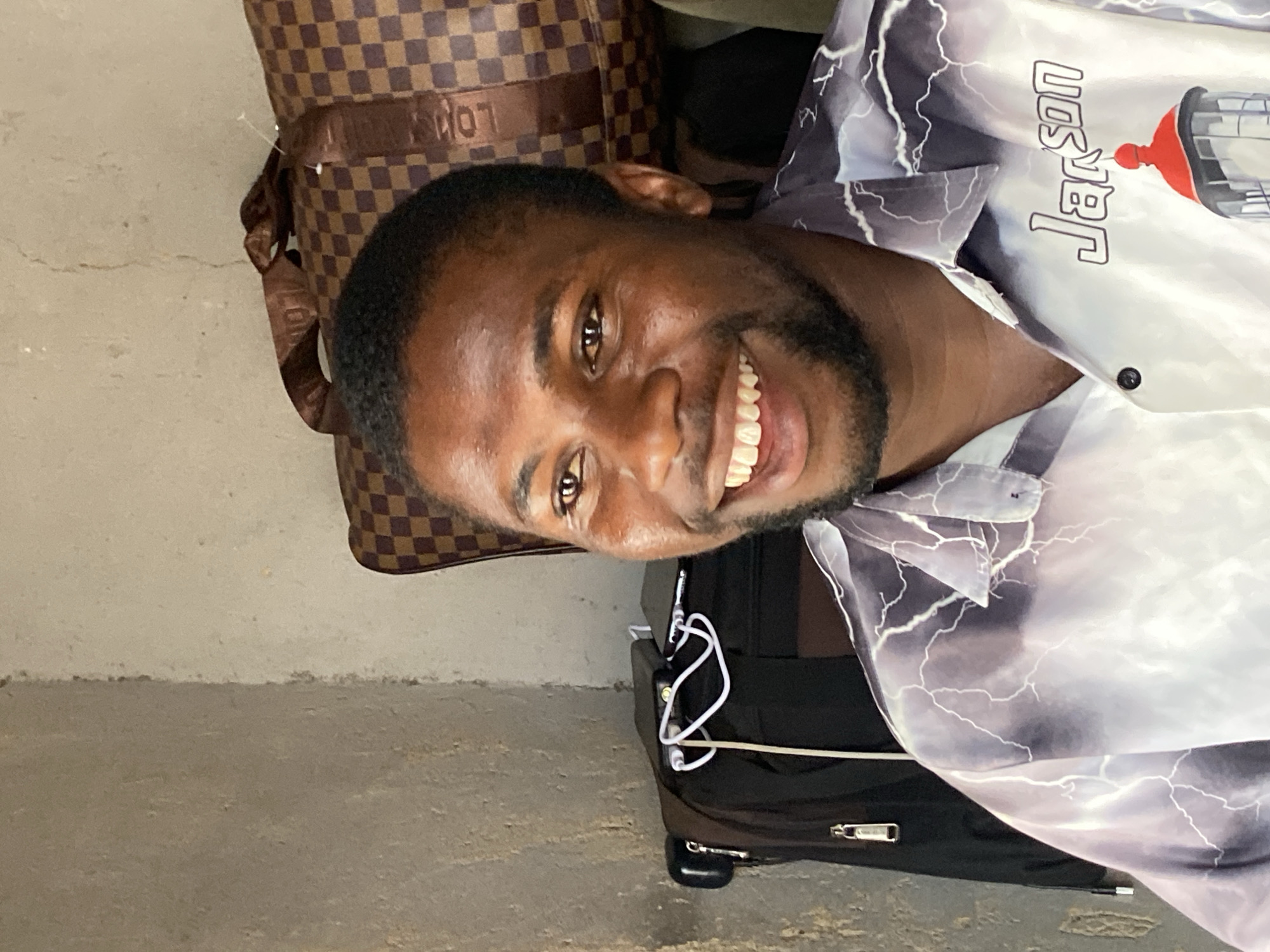 The image captures a close-up of a man smiling broadly and looking directly at the camera, likely posing for a photograph in Bukuru, Nigeria. He has dark skin, short dark hair, and a well-groomed beard, with a wide, joyful smile revealing his white teeth.
The man is wearing a button-up shirt featuring a distinctive pattern of white lightning bolts against a gray and purple background. On the right side of his collar, the word "Jarson" is prominently displayed, next to a graphic that appears to be a stylized lighthouse with a red top.
Behind the man's head and to his left, a brown and tan checkered bag or suitcase is visible, featuring a partially visible and inverted logo that resembles "LOUIS". To his right and below, a black rolling suitcase can be seen, with white wires, possibly from headphones or a charging cable, draped over it.
The setting appears to be indoors or in a covered area, with a light-colored, textured wall forming the background, showing some minor cracks or imperfections. The floor is a light gray, textured surface, possibly concrete. The man's bright smile conveys a friendly and happy emotion.